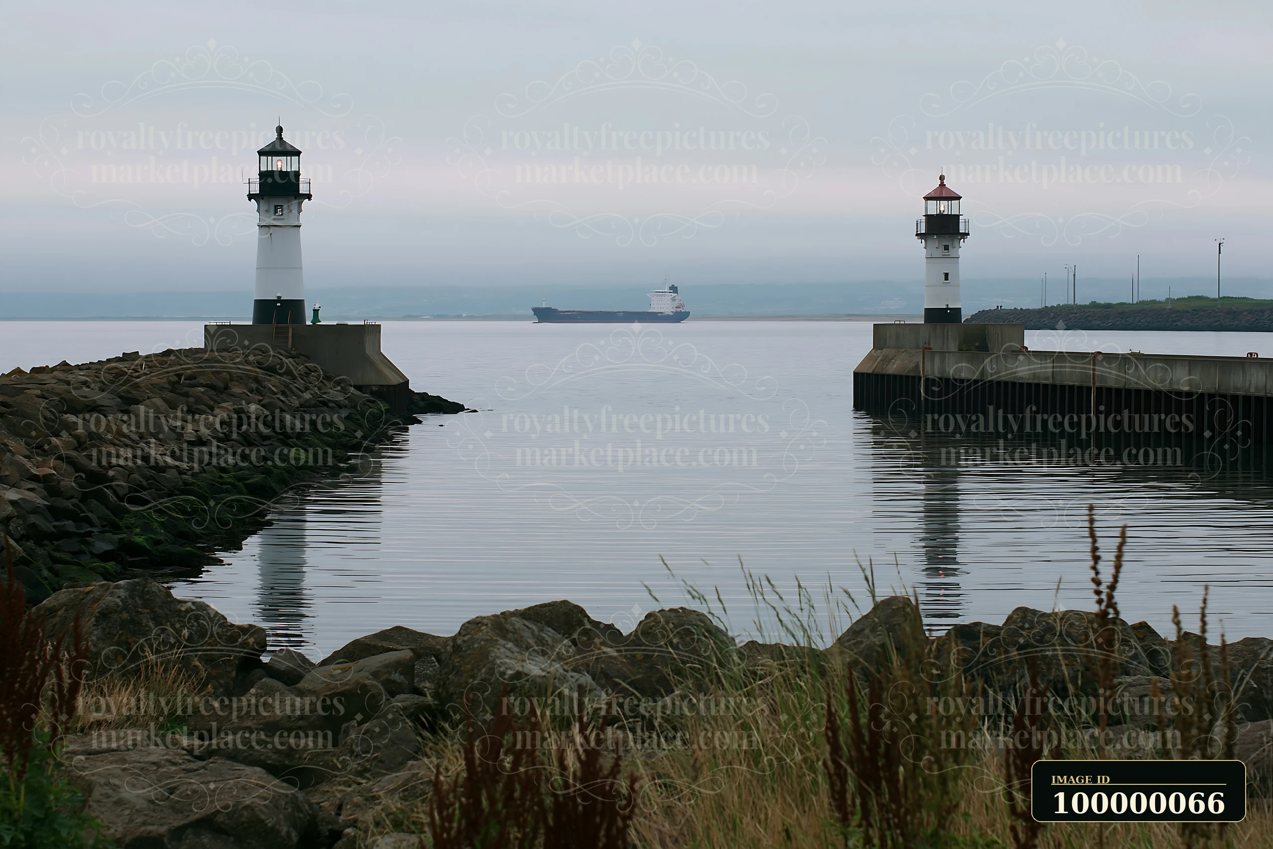 Twin harbor lighthouses frame a cargo ship sailing across calm water beneath an overcast sky.