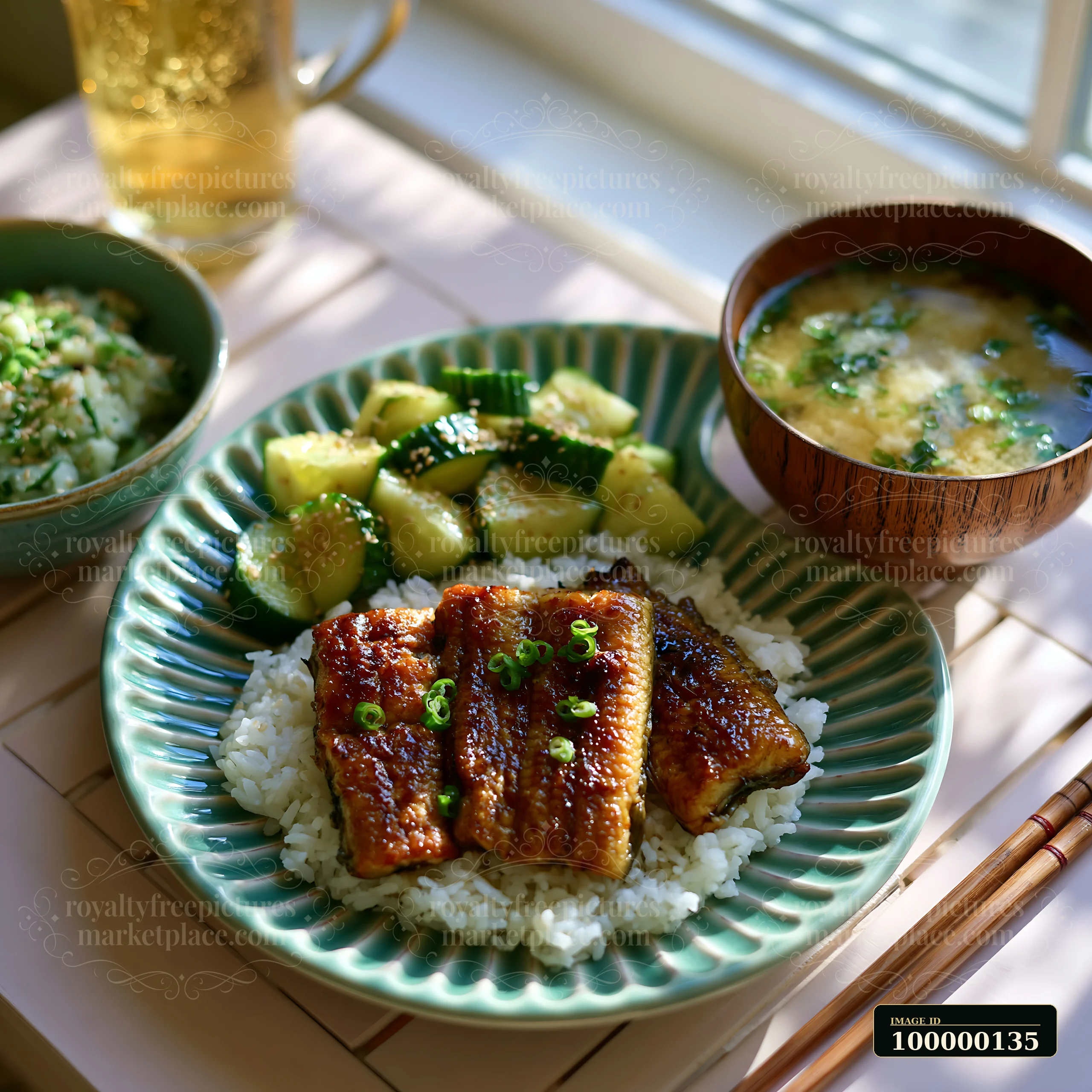 Japanese breakfast plate with glazed fish over rice, vegetables, soup, tea, and chopsticks on a sunlit table.