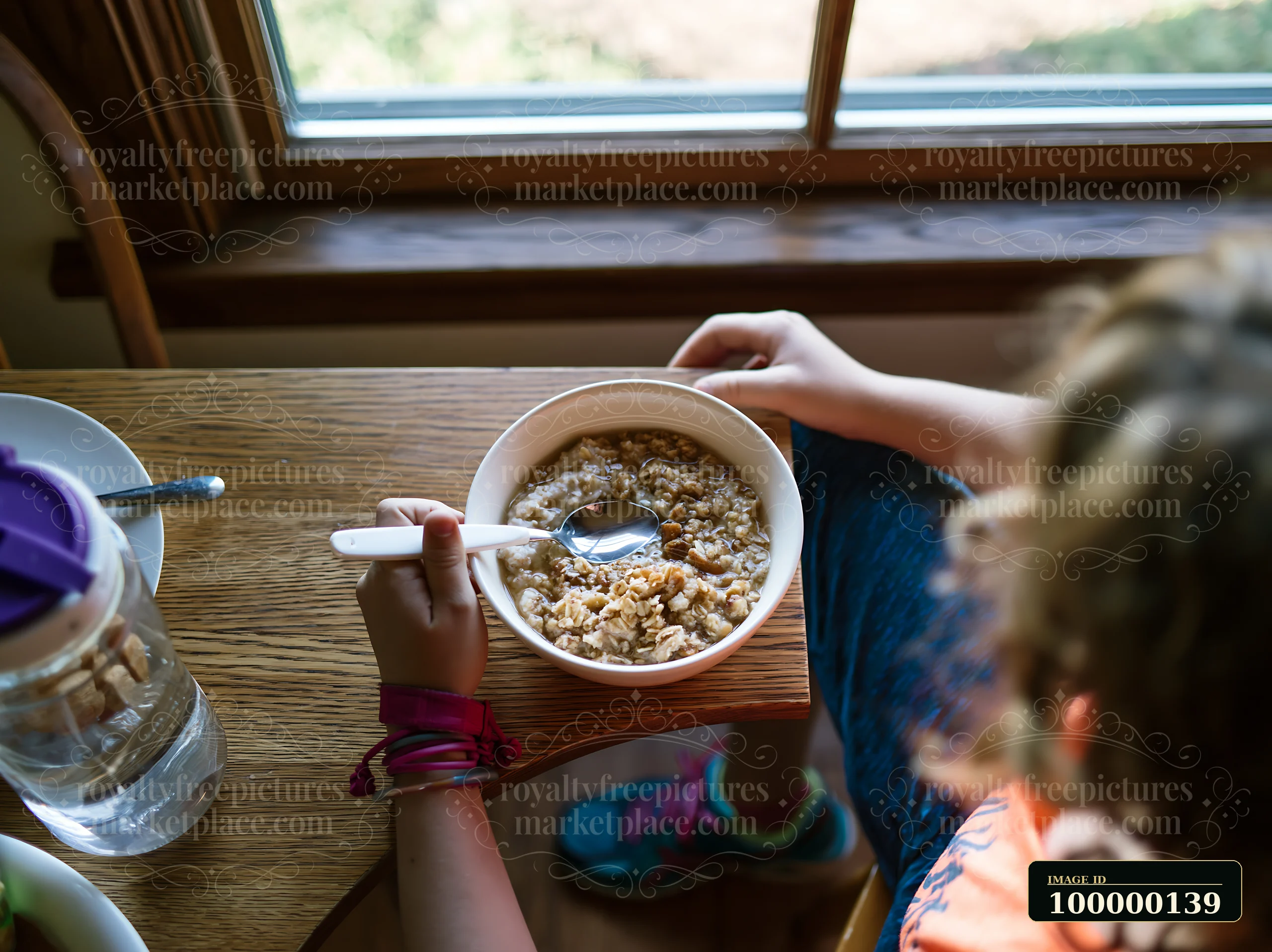 Person uses an adapted spoon to eat cereal at a wooden table beside a sunlit window.