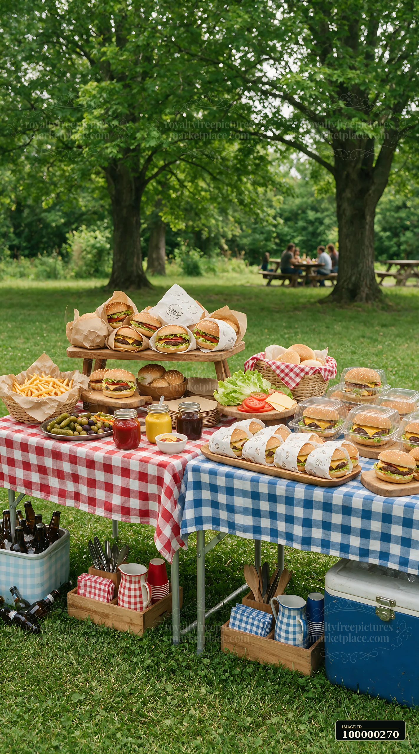 Outdoor burger buffet with burgers, fries, condiments, vegetables, and picnic tables beneath leafy trees.