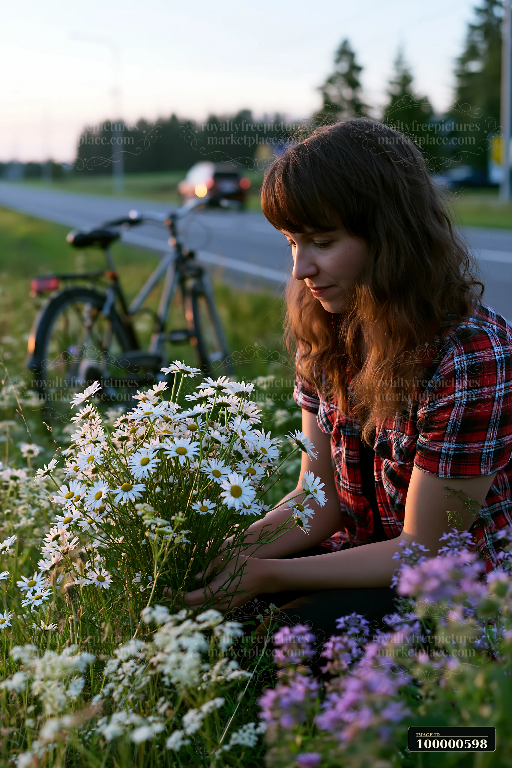A woman sits on the grass near the road, gathering flowers while a bicycle leans beside her.
