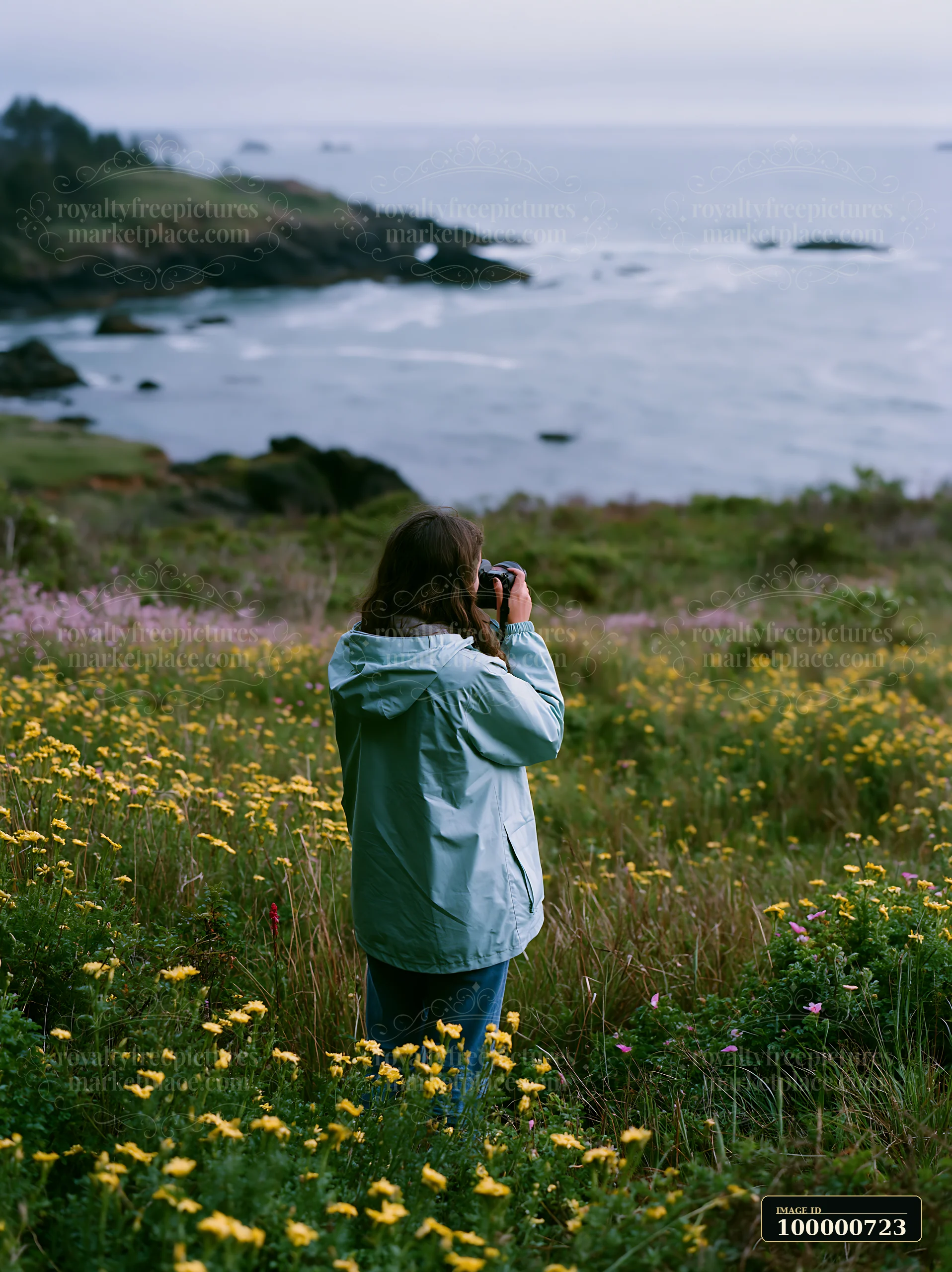 A woman stands in a coastal meadow overlooking ocean, surrounded by colorful flowers and grass.