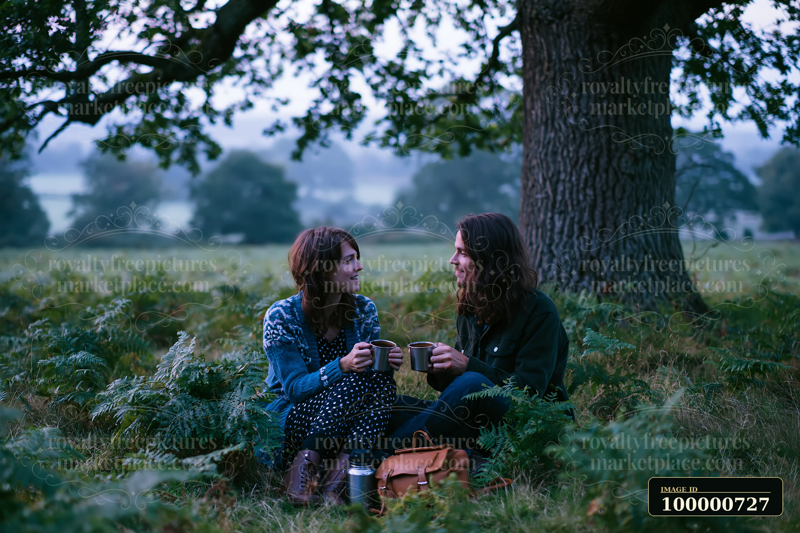 Two people sit in a meadow beneath an oak tree, holding cups and sharing a moment together.
