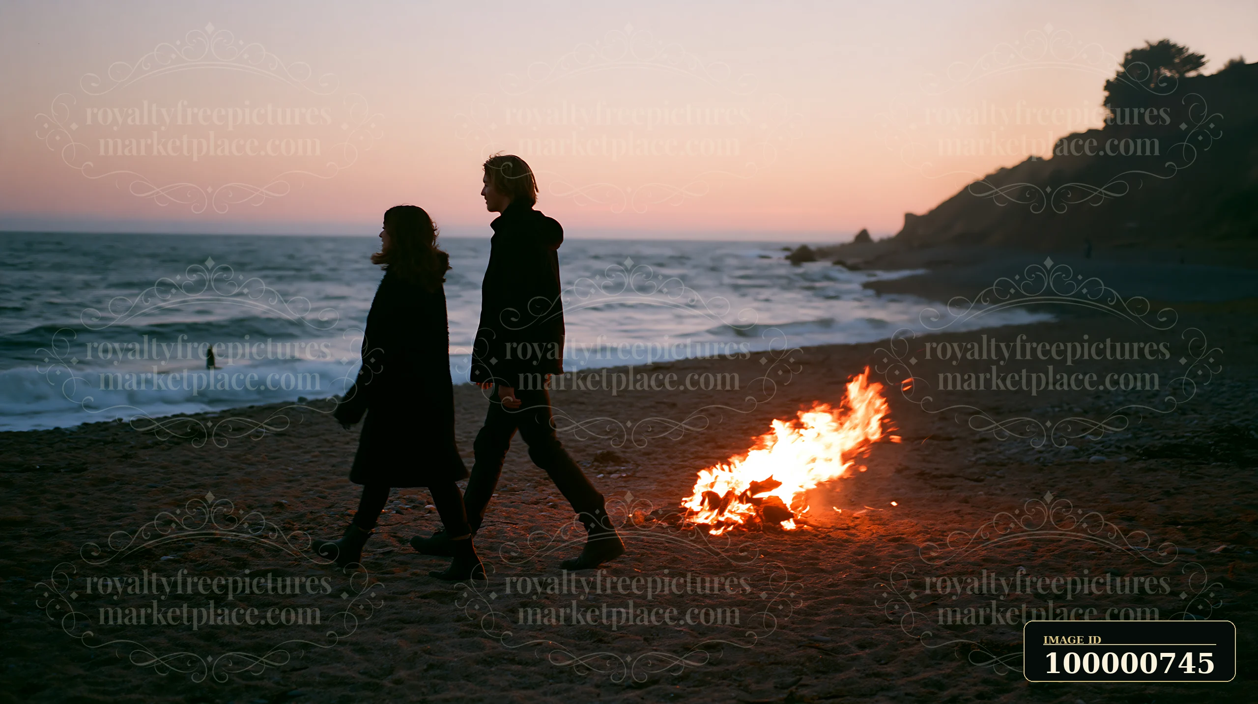 Two individuals walk along a sandy beach at sunset, with waves crashing nearby and a fire burning on the sand.