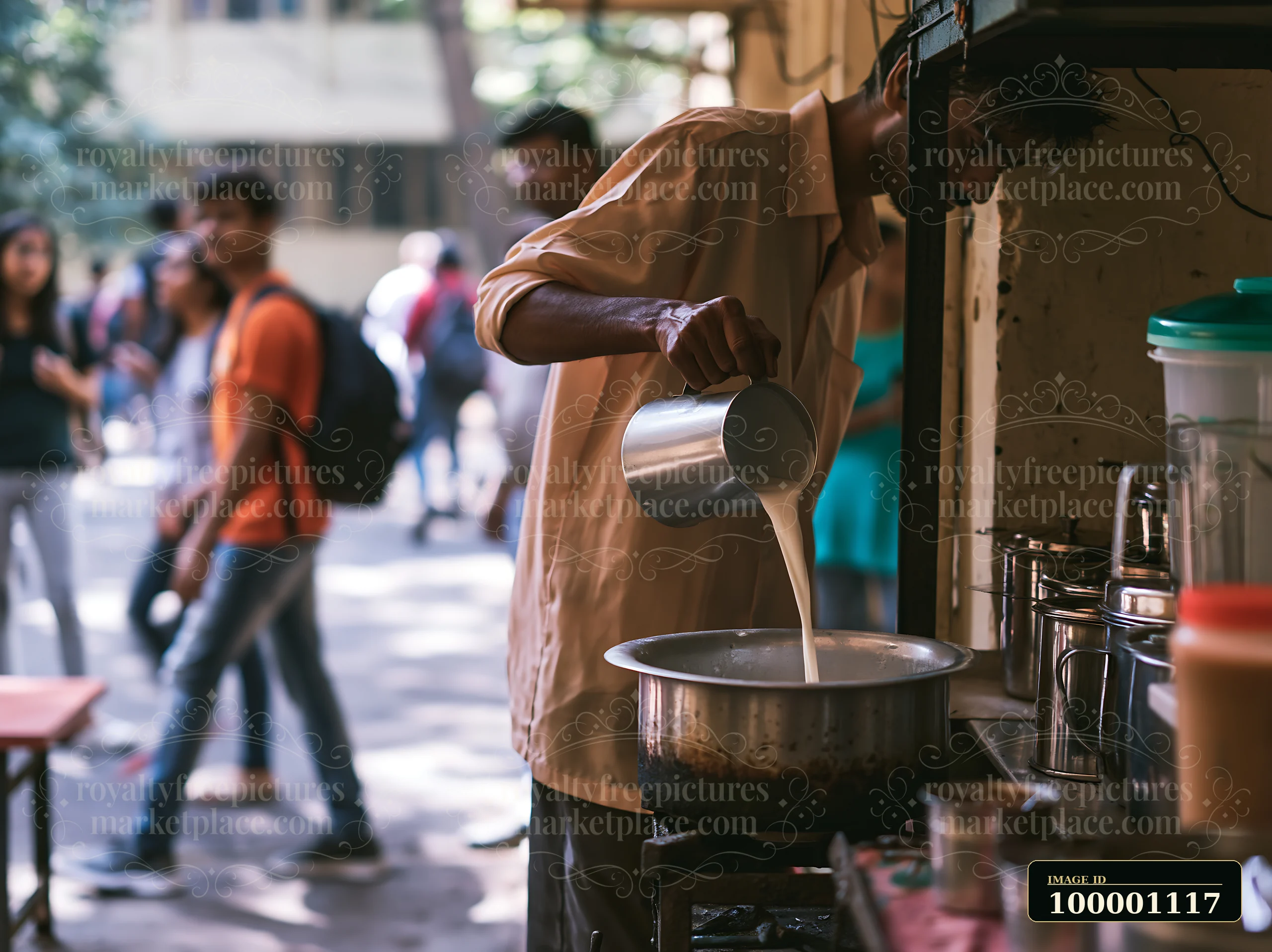 A street food vendor is pouring milk into a pot of tea at a bustling outdoor setting with people walking by.