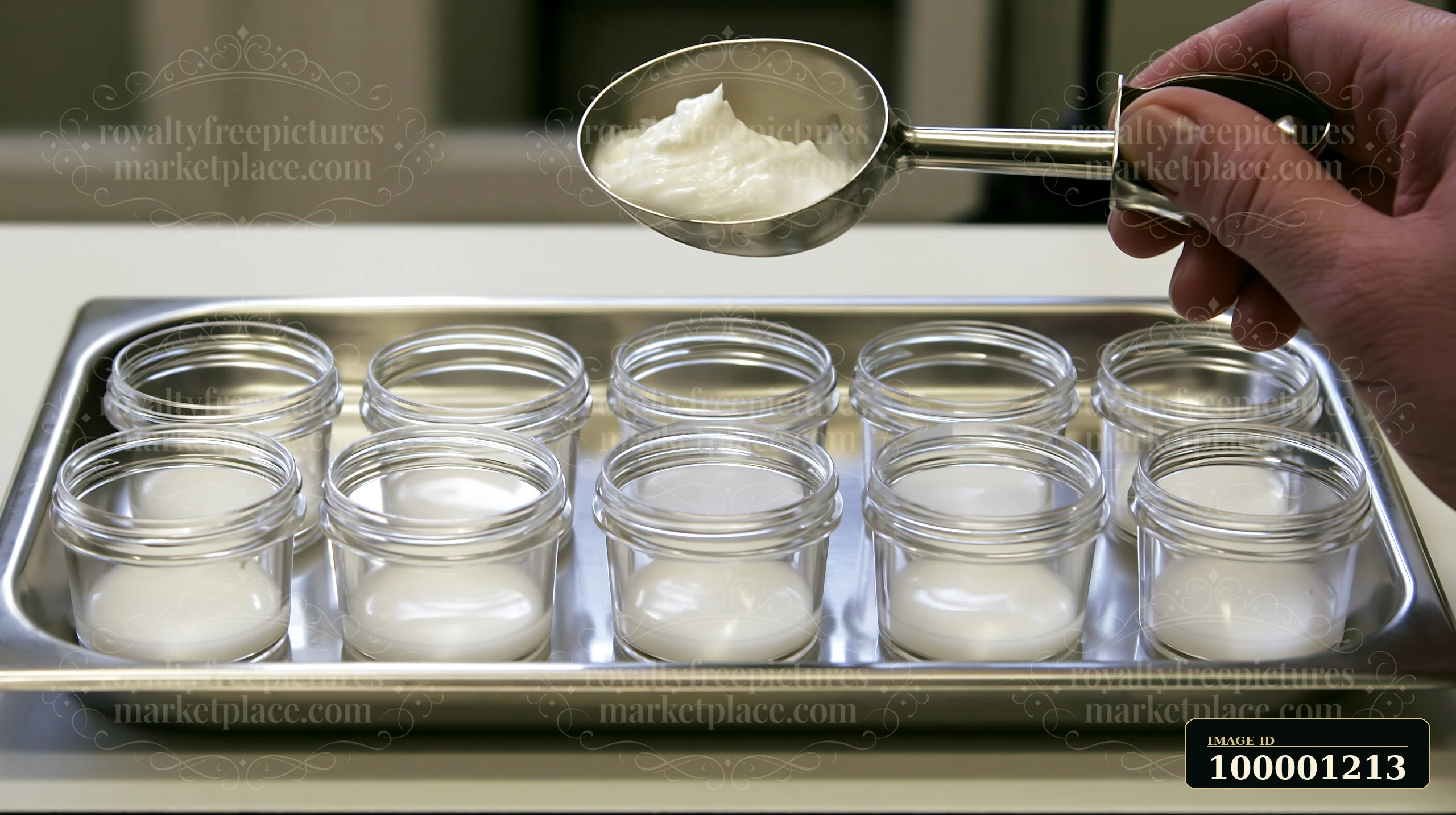 A person holds a metal scoop filled with cream above several small glass jars on a stainless steel tray.