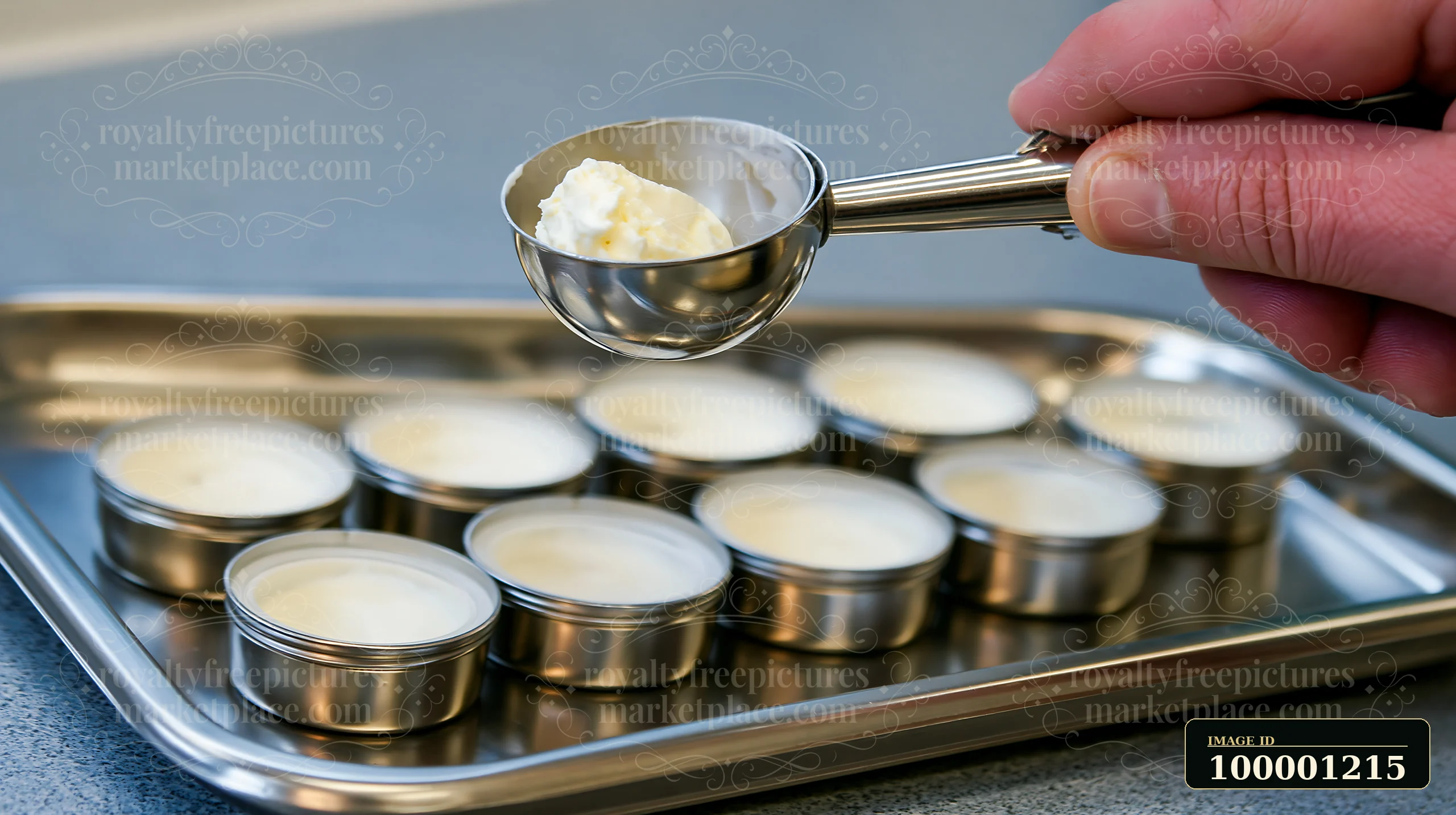 A hand holds a metal scoop measuring cream over a tray filled with small containers.
