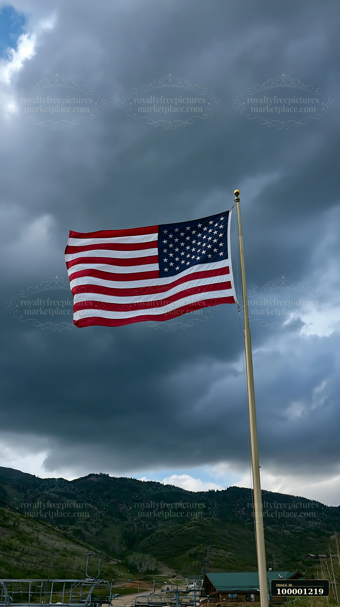 The American flag flying in strong wind atop a flagpole against a backdrop of dark clouds and rolling hills.