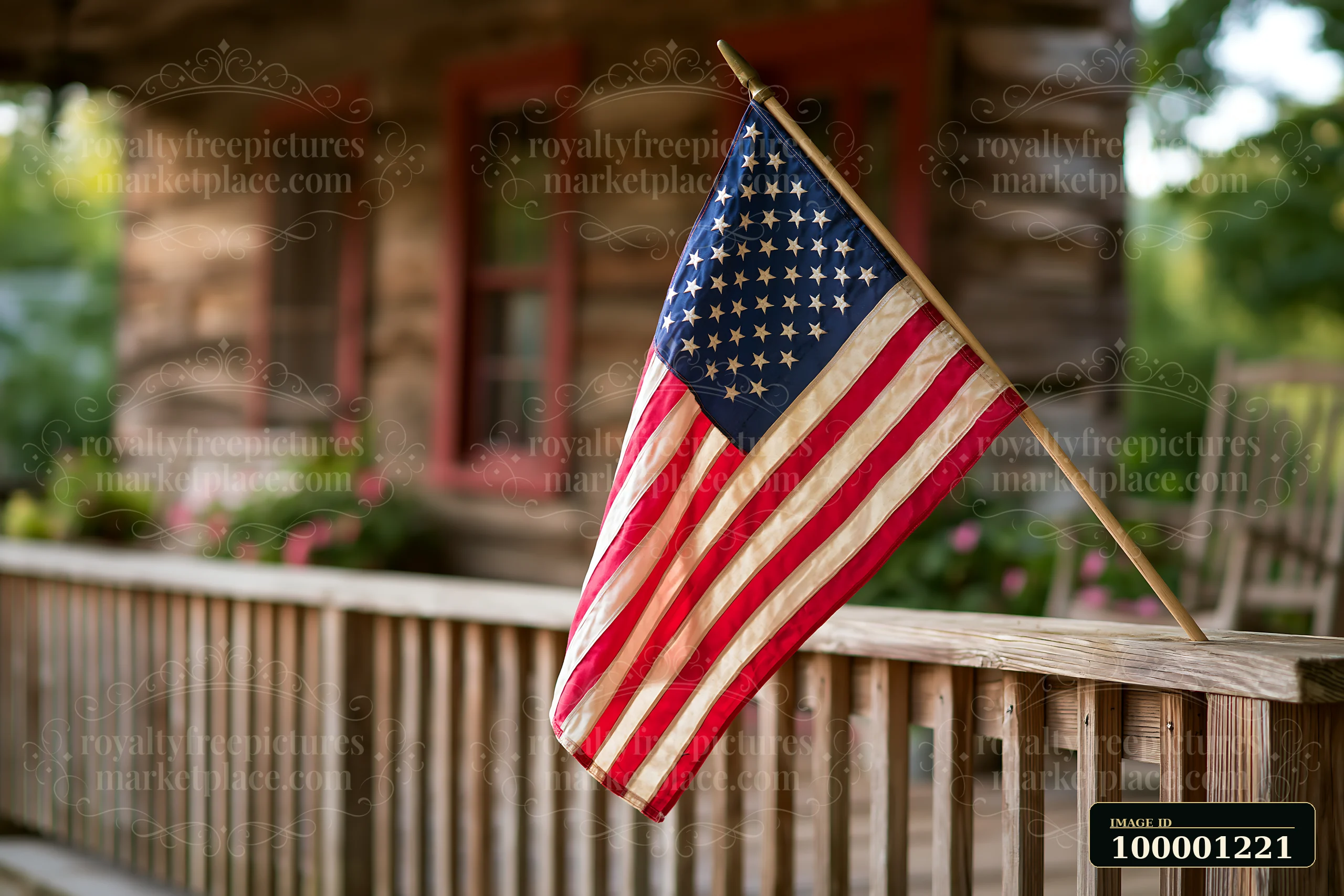 The American flag waving in gentle breeze hangs from a wooden pole beside a rustic house with red-framed windows and a wooden porch filled with blooming flowers