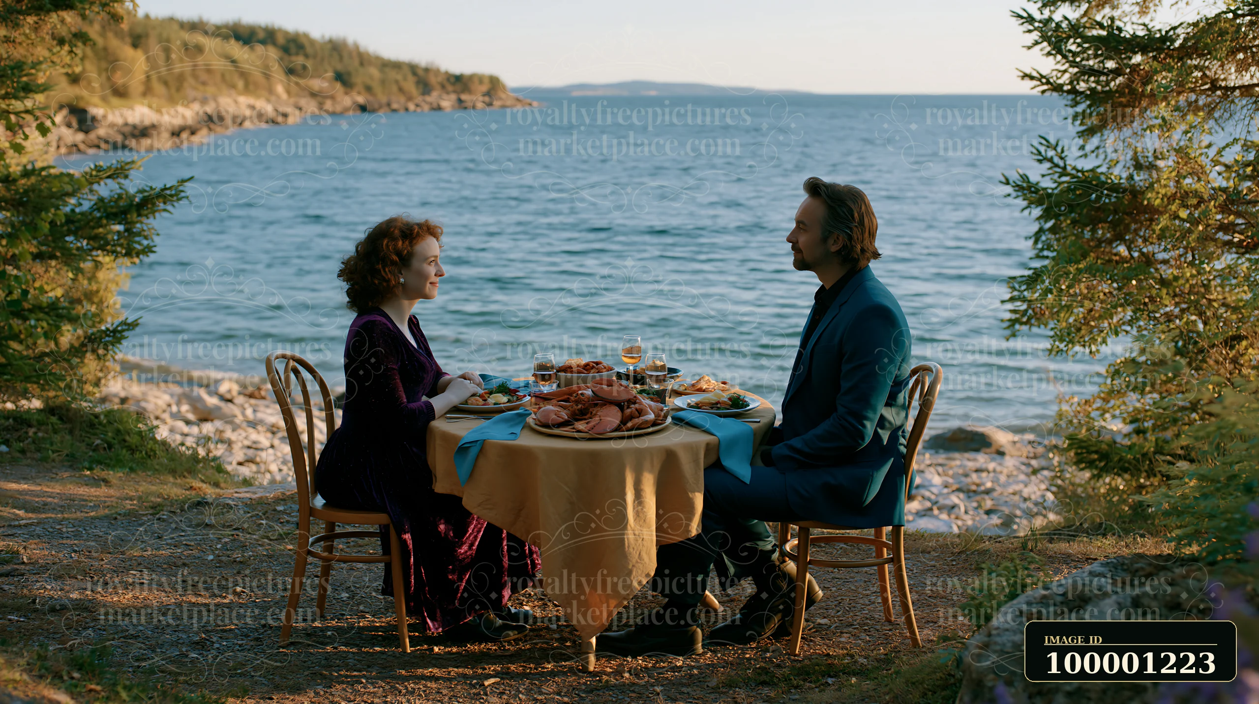 A couple sits at a table set for a picnic buffet by the water, surrounded by trees and rocks.