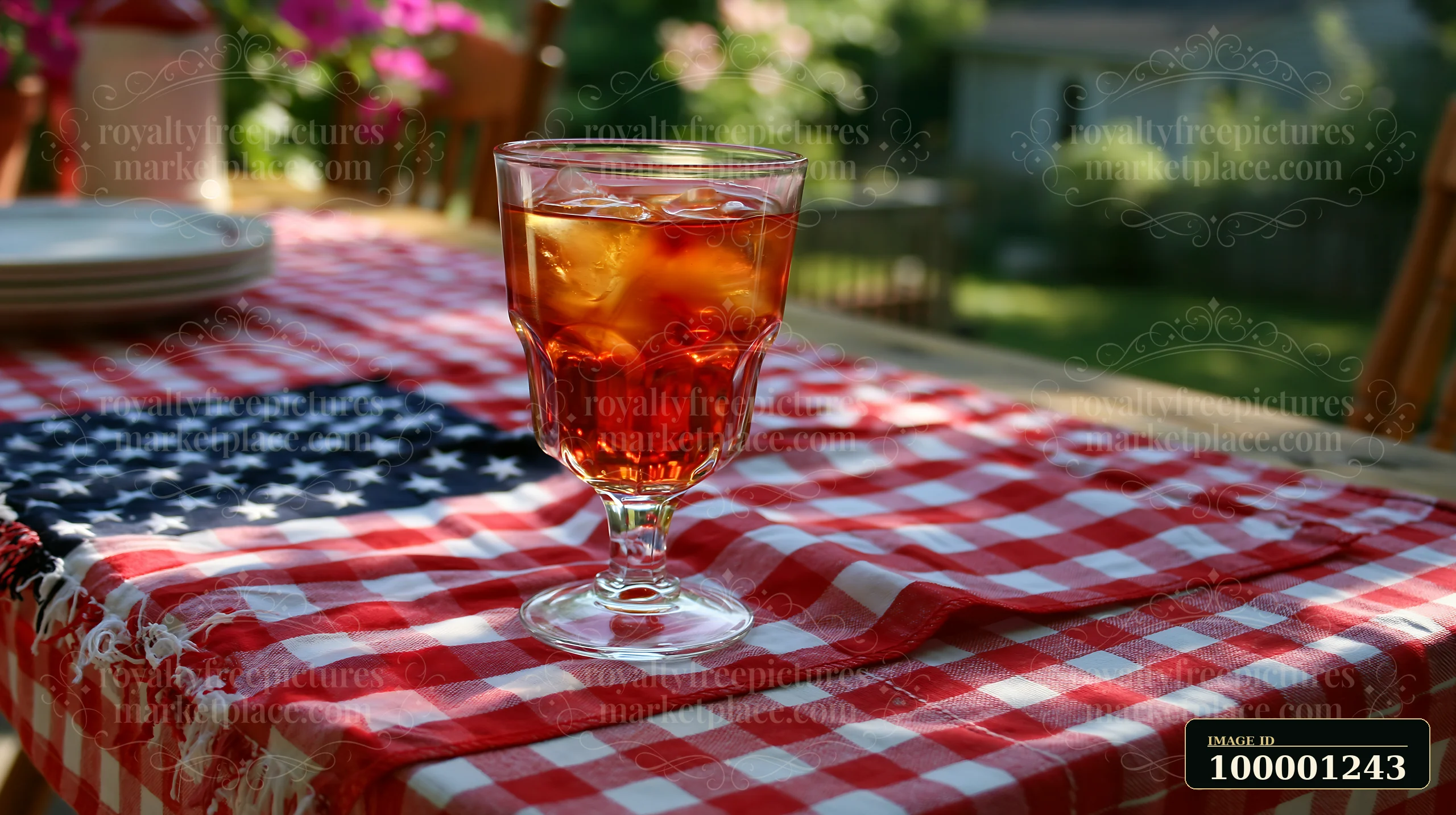 A glass filled with iced tea sits on a table covered with an American flag table decoration and a red and white checkered cloth in a sunny outdoor setting.