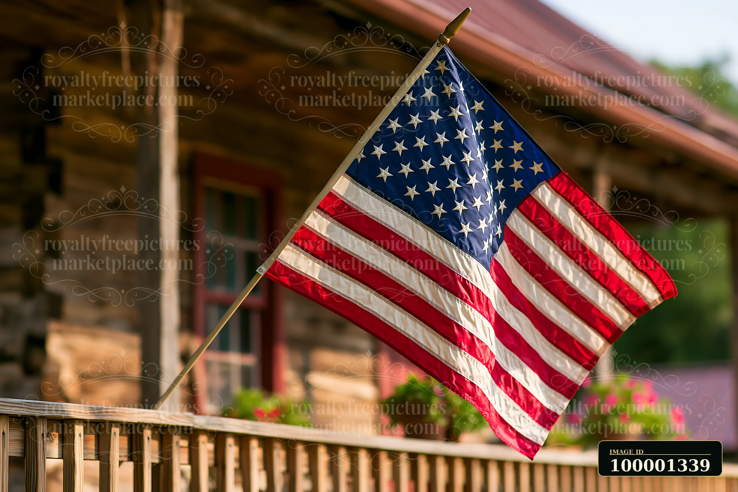 An American flag waving in gentle breeze is displayed on a porch of a wooden cabin surrounded by potted flowers.