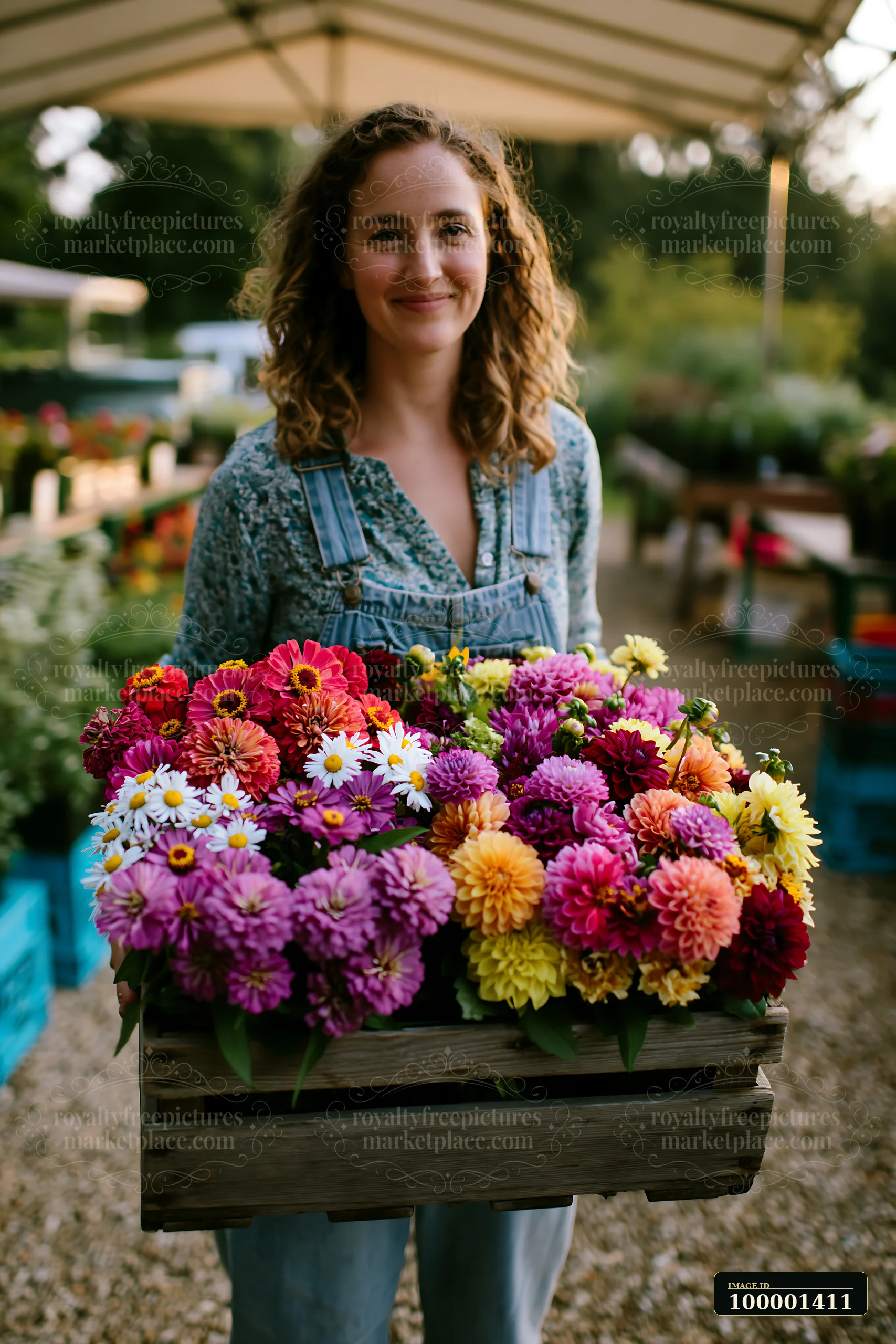 A woman holds a wooden crate filled with colorful mixed seasonal flowers at a farmers market.