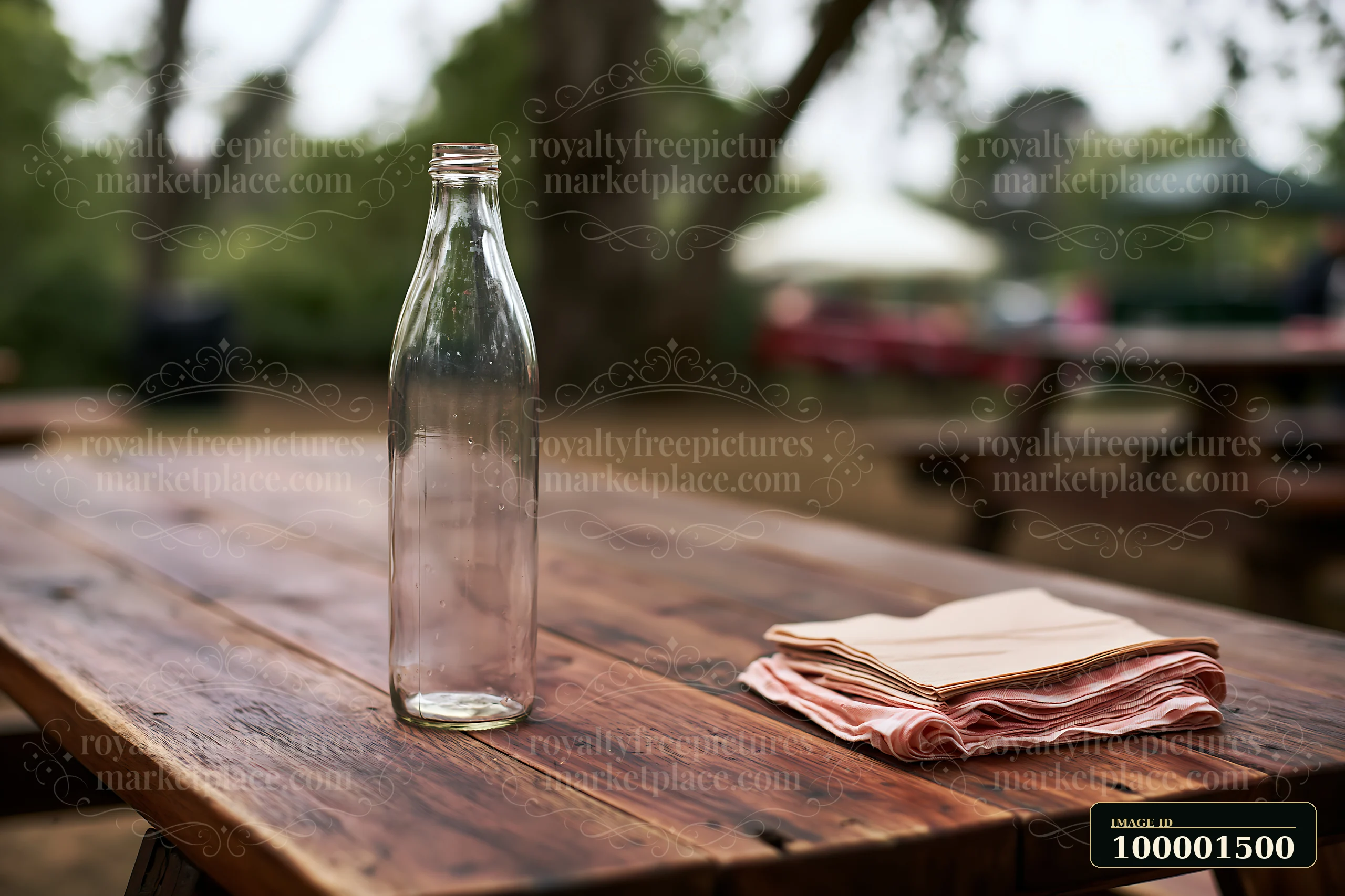 An empty bottle is placed on a wooden table beside folded napkins at an outdoor event, surrounded by trees and distant picnic tables.