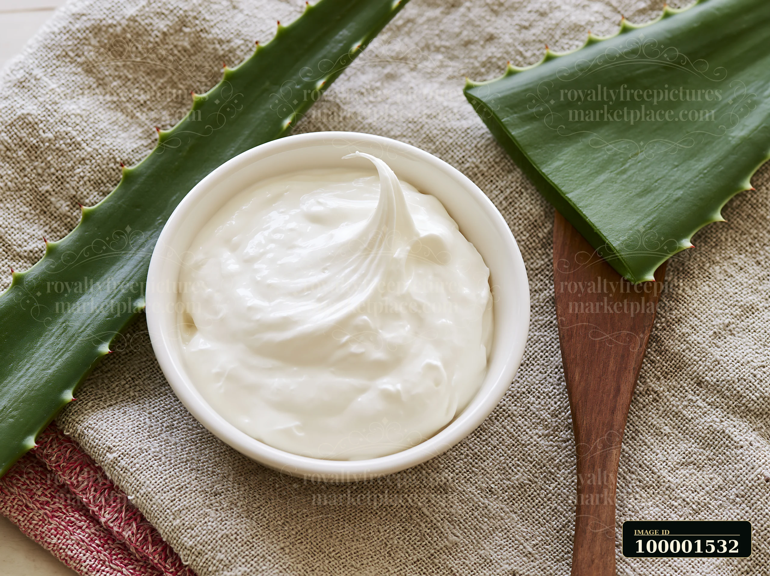 A bowl of cream sits next to fresh aloe vera leaves on a textured cloth.