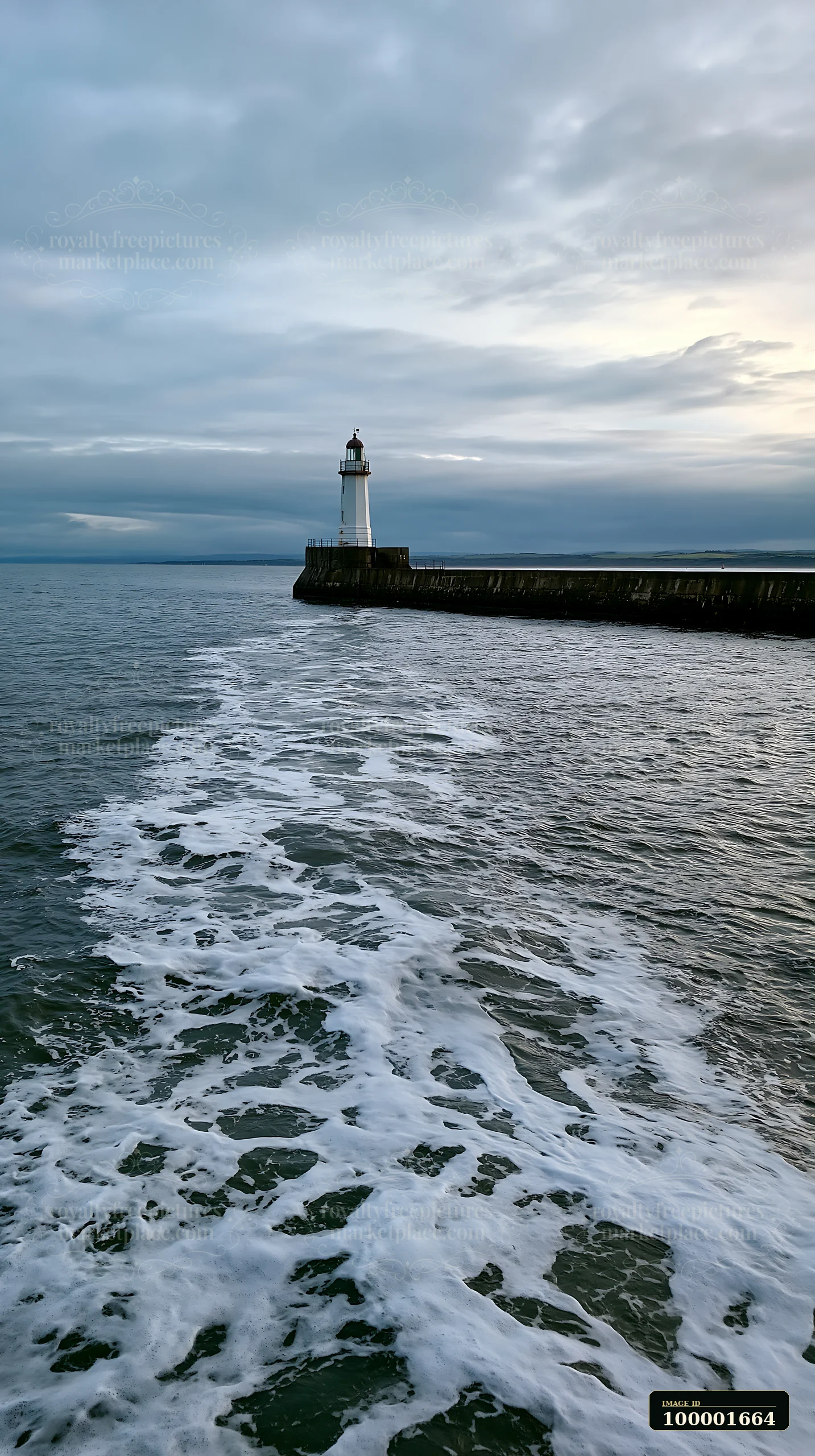 A lighthouse stands on a rocky pier as waves splash against it under a cloudy sky at sunset.