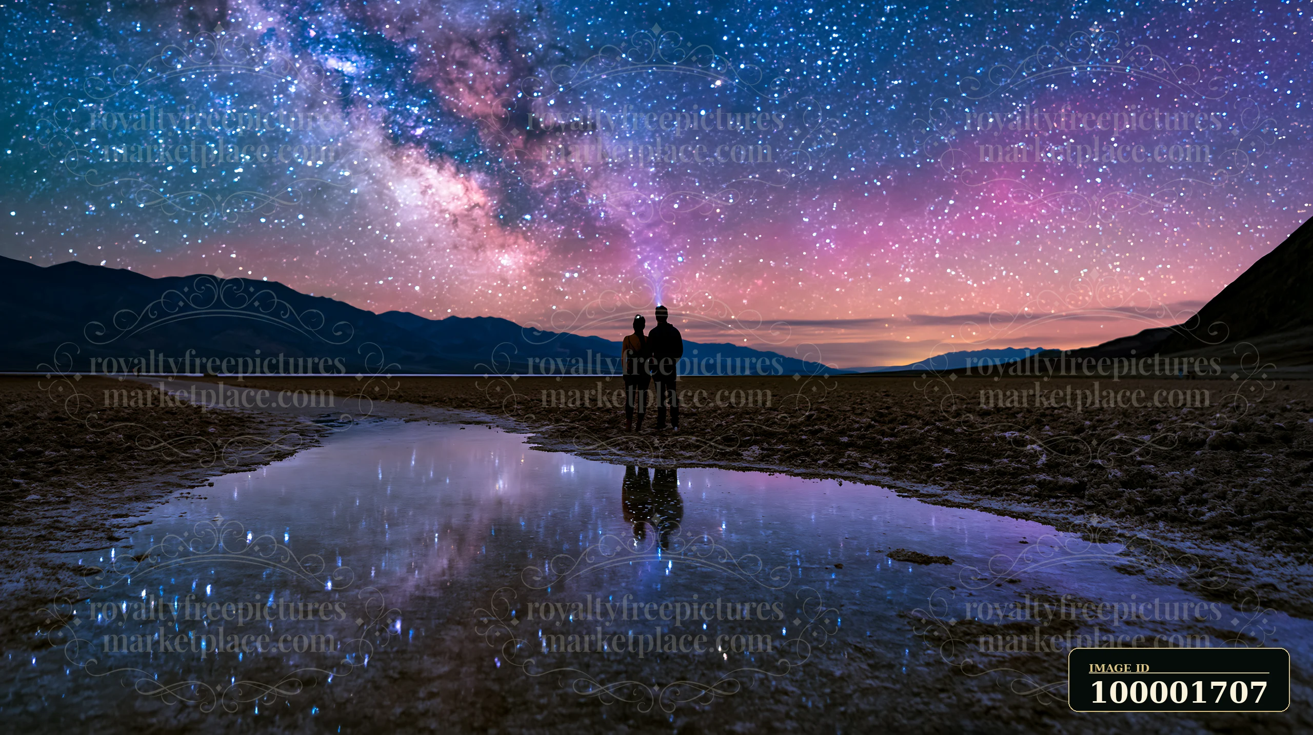 A couple stargazing in a desert at night stands near a body of water reflecting the Milky Way.
