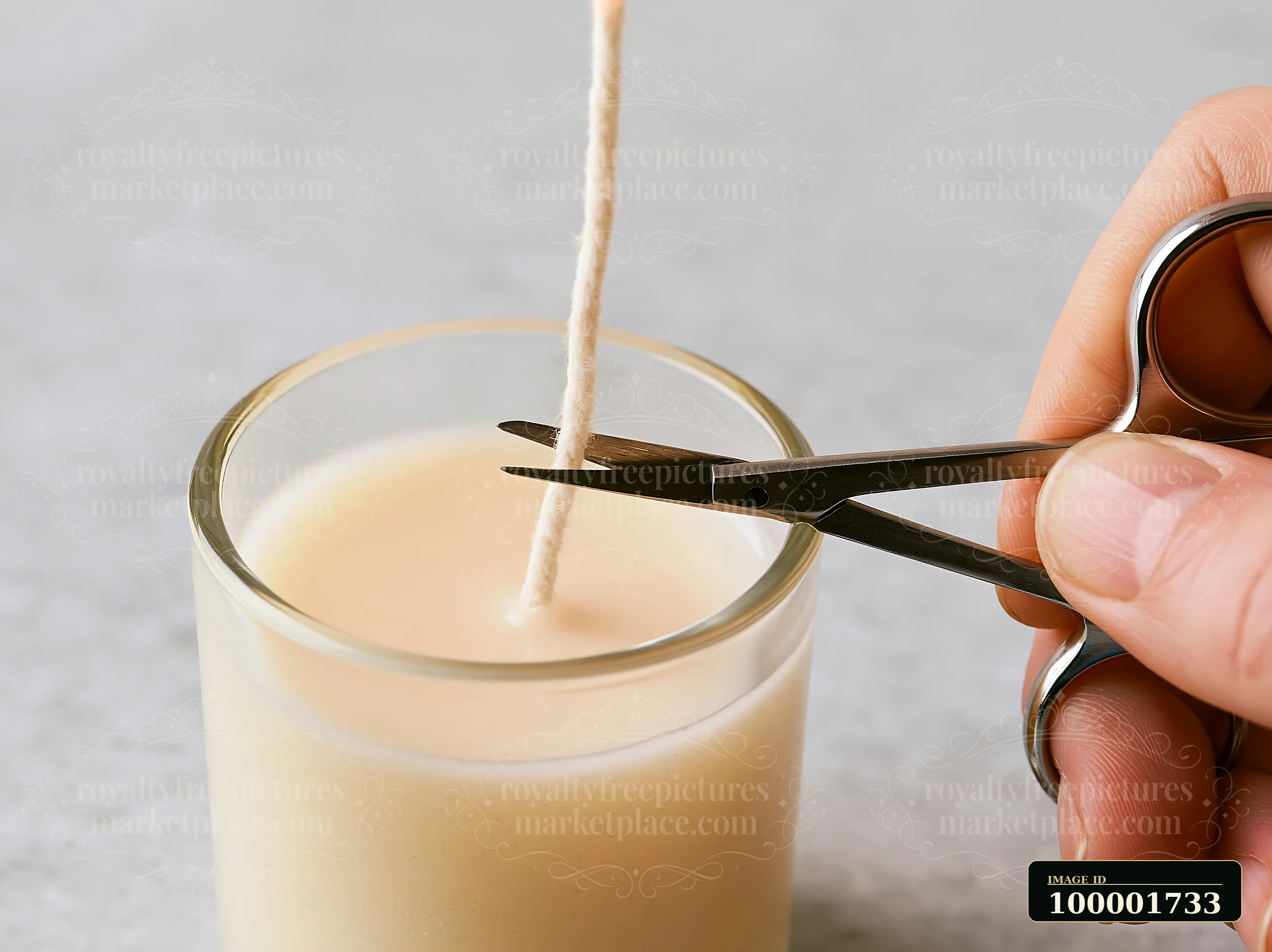 A hand uses scissors to trim the wick of a vanilla candle in a clear glass container against a neutral background.