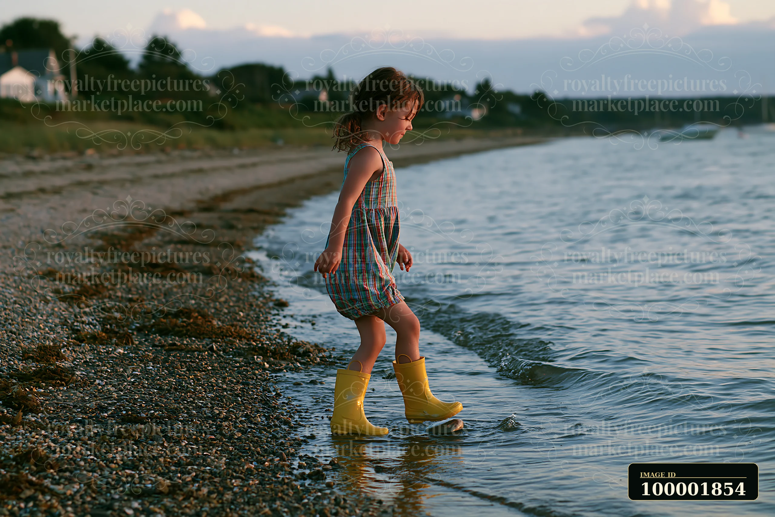 A child in yellow rain boots walks along a pebbly coastline beach, splashing in the water during sunset.