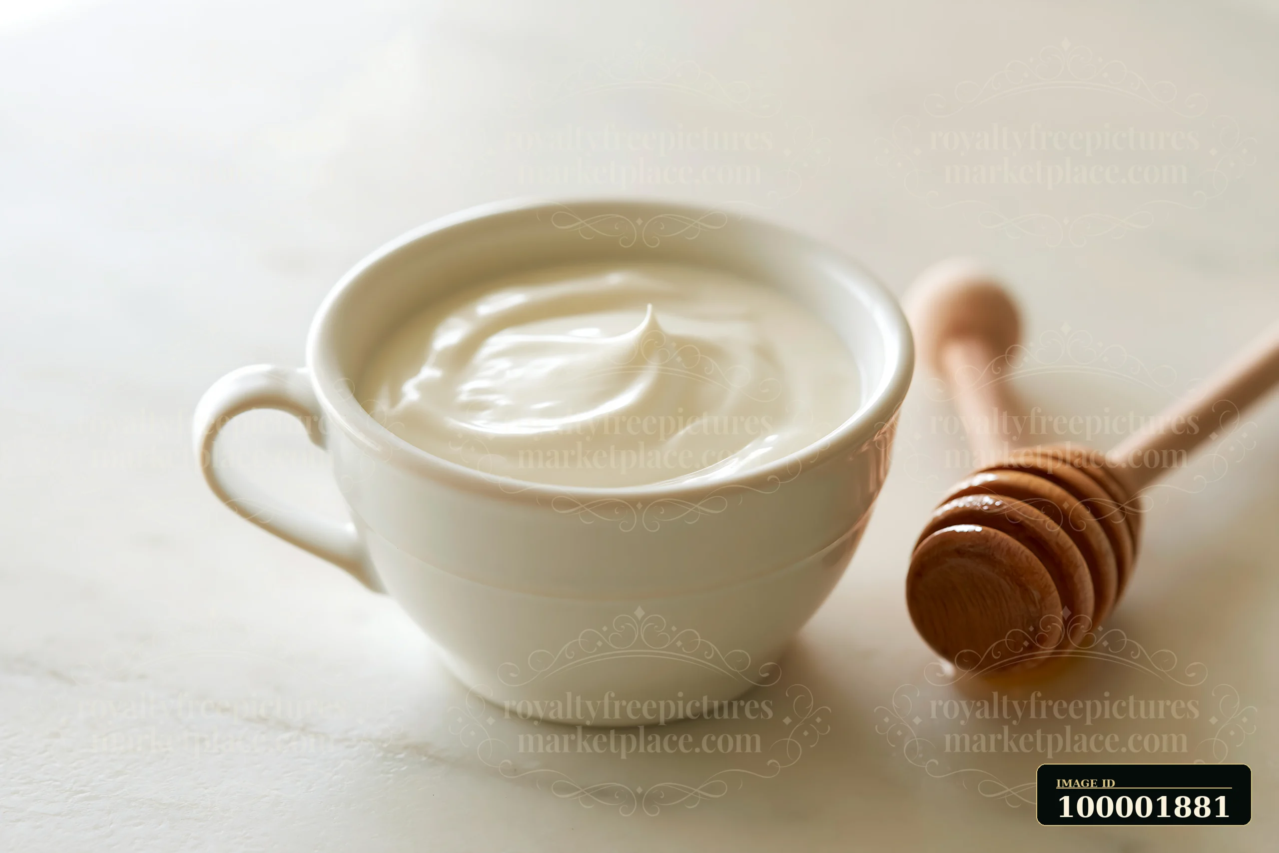 A cup filled with cream sits next to a wooden honey dipper on a light surface.