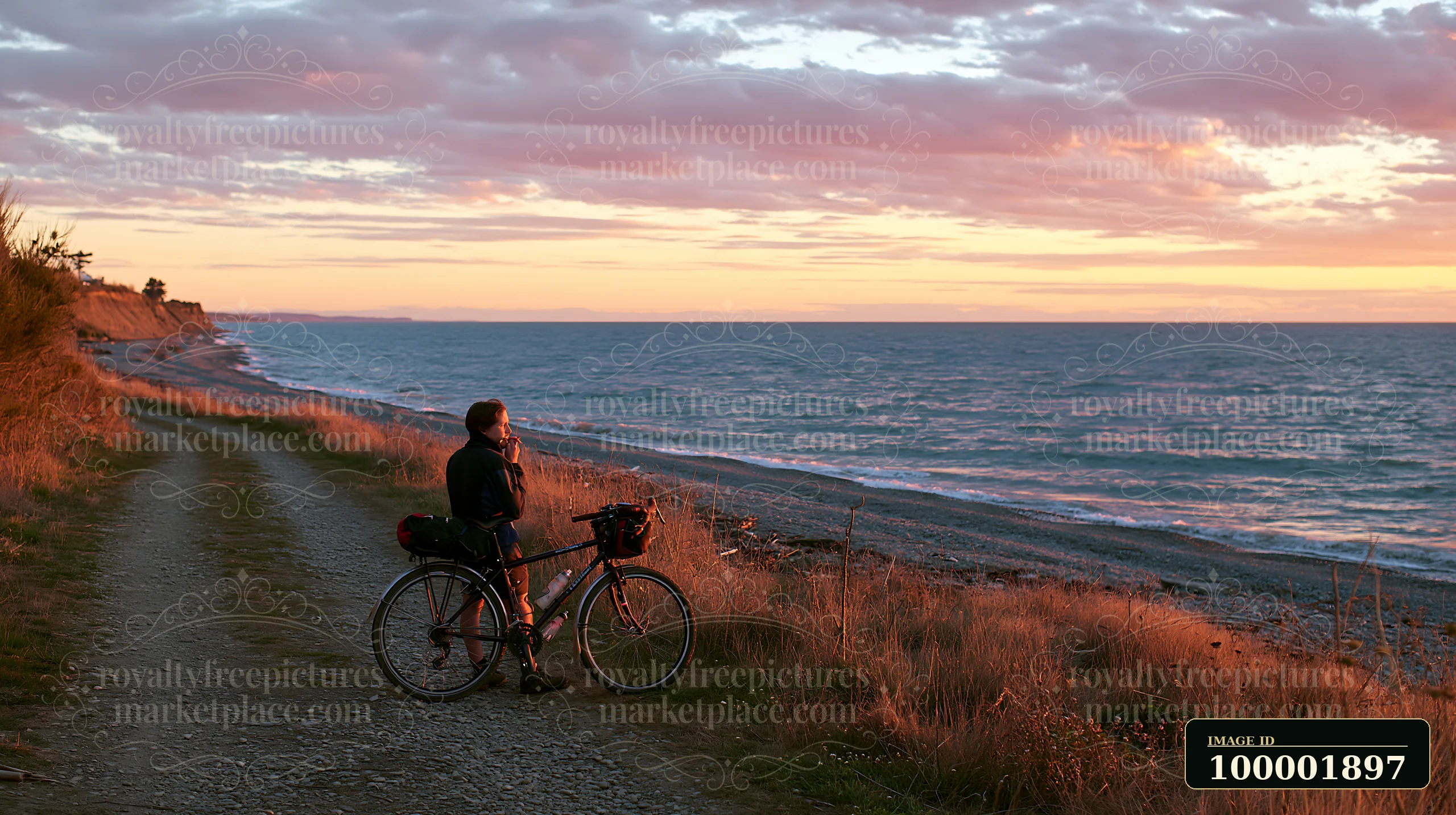 A person stands beside a bicycle on a dirt path, watching the sunset over the pebbly coastline beach.