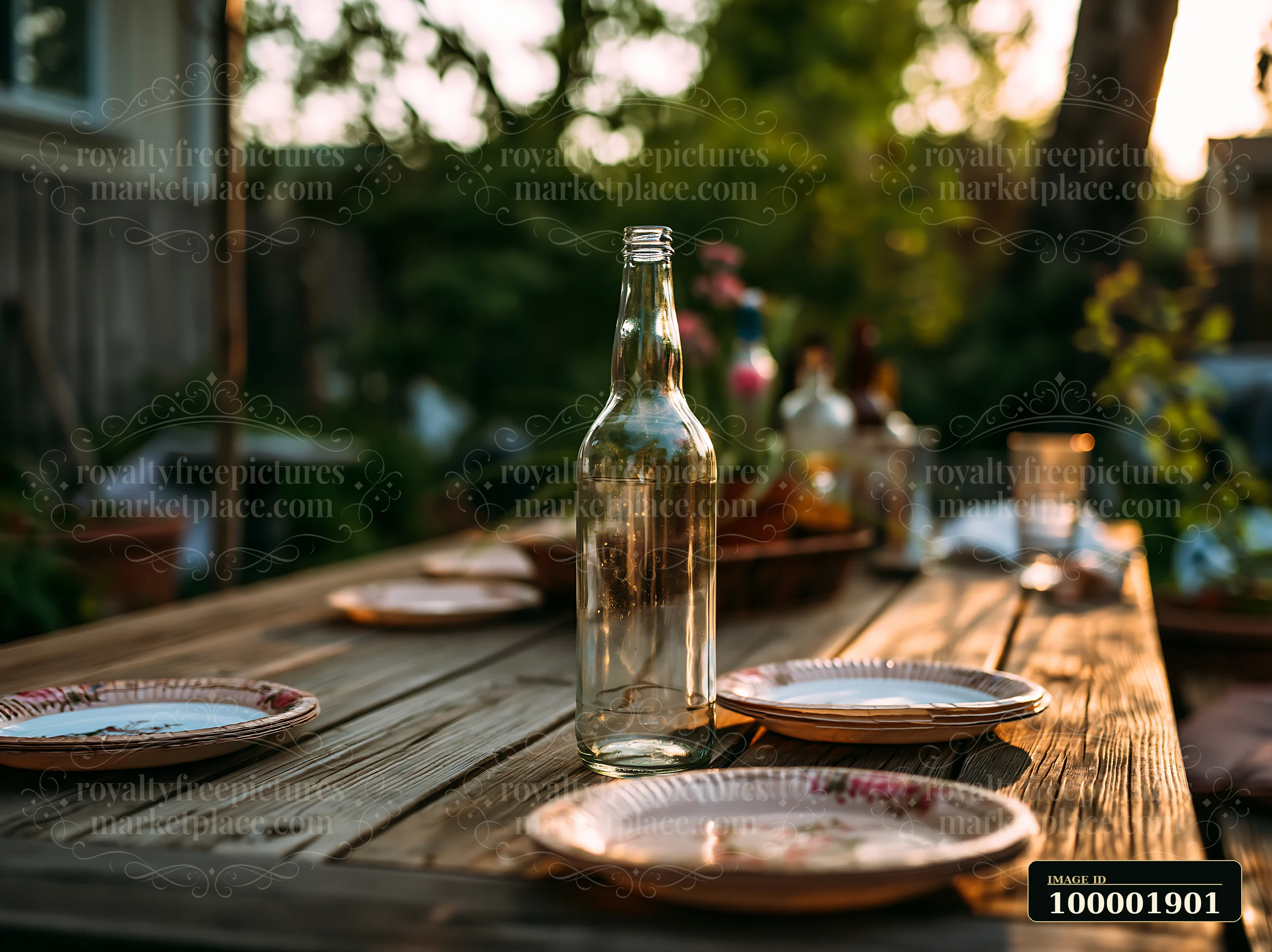 An empty bottle stands on a wooden table surrounded by used plates at a backyard gathering as the sun sets, creating a relaxed atmosphere.