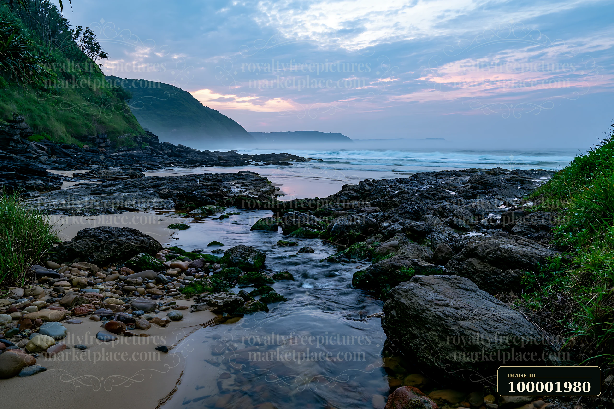 The coastline with exposed rocks features a stream flowing through the area, surrounded by grass and sand as waves lap gently at the shore during twilight.