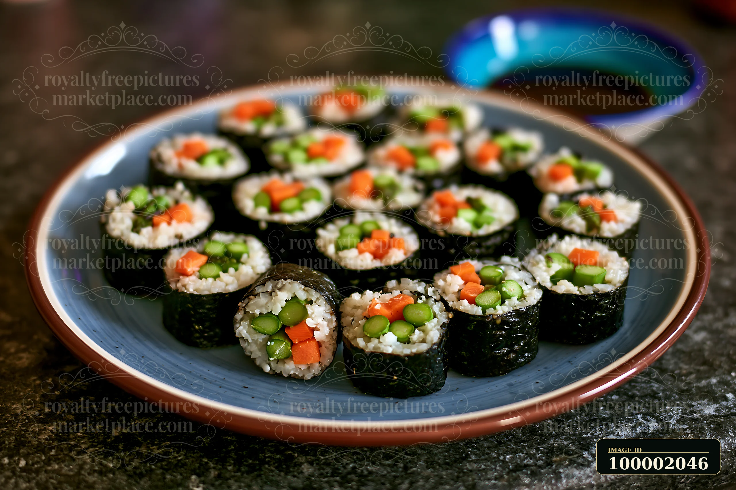 A vegan sushi platter features rolls made with rice, nori, carrots, and green vegetables arranged neatly on a blue patterned plate.