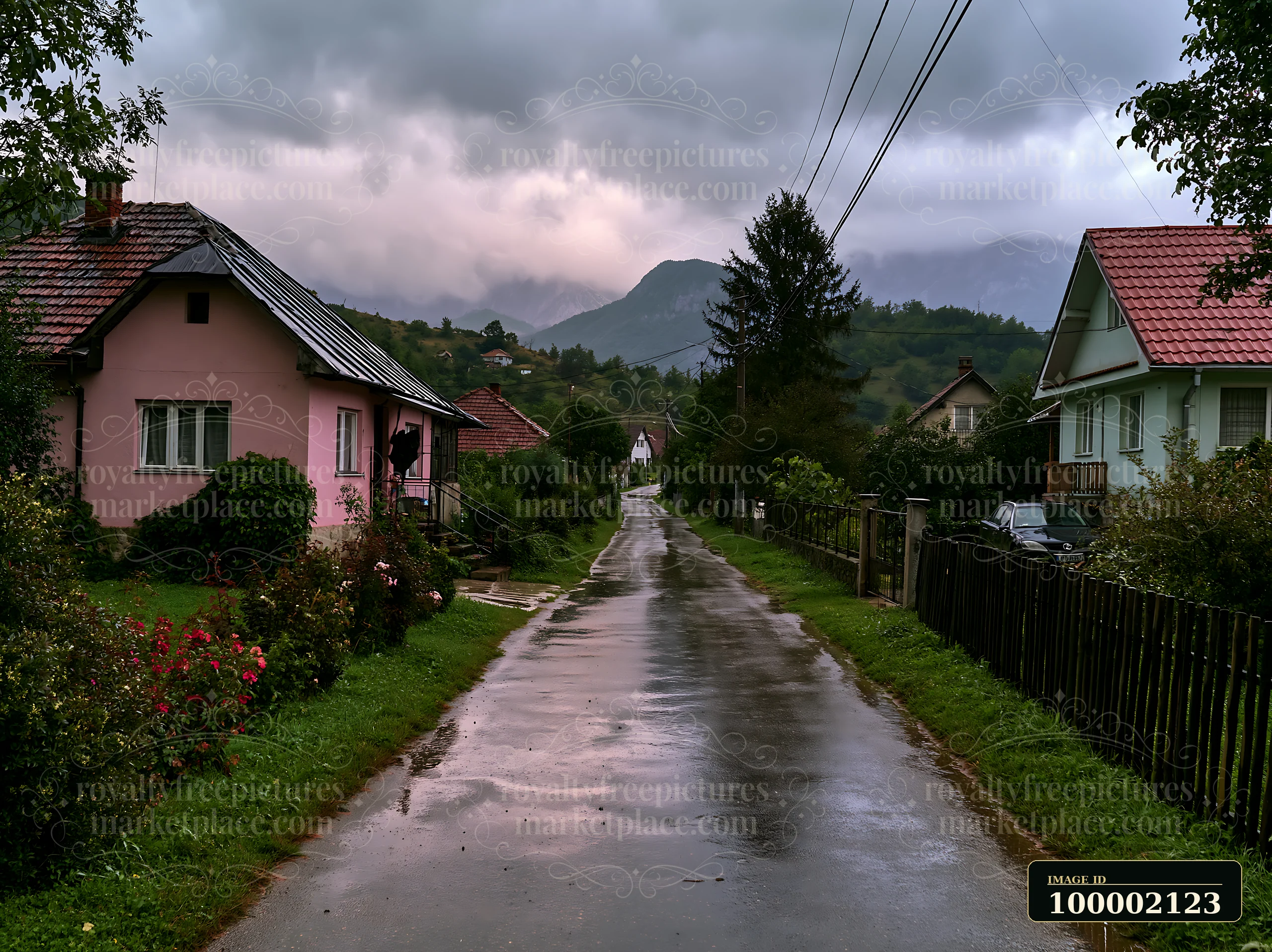 Dark clouds gather over the mountain range as rain falls on a quiet rural road lined with houses.