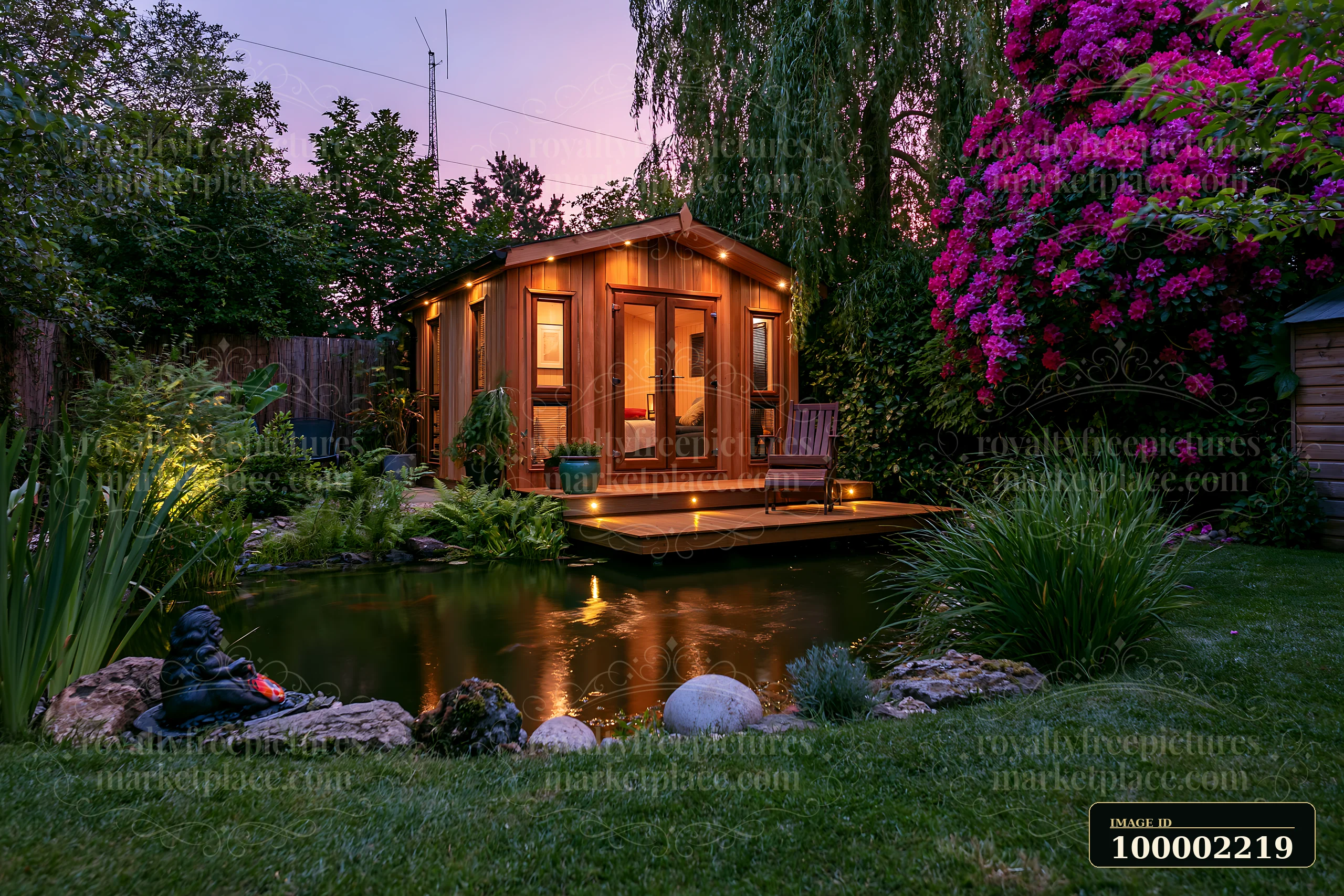 A garden room with an illuminated deck sits beside a pond, surrounded by flowers and plants.