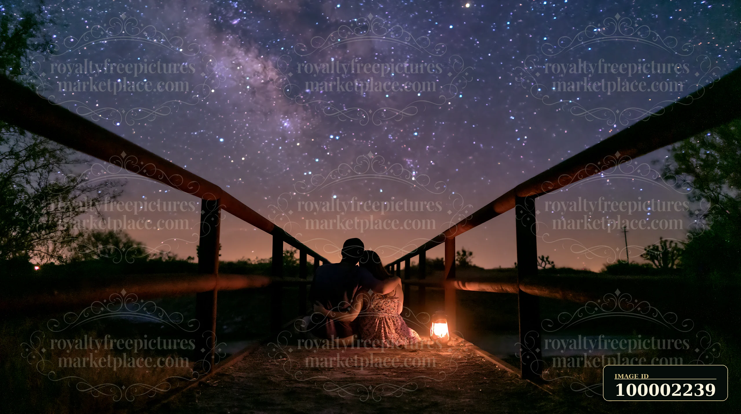 A couple stargazing in a desert at night sits on a wooden bridge under a clear sky filled with stars.