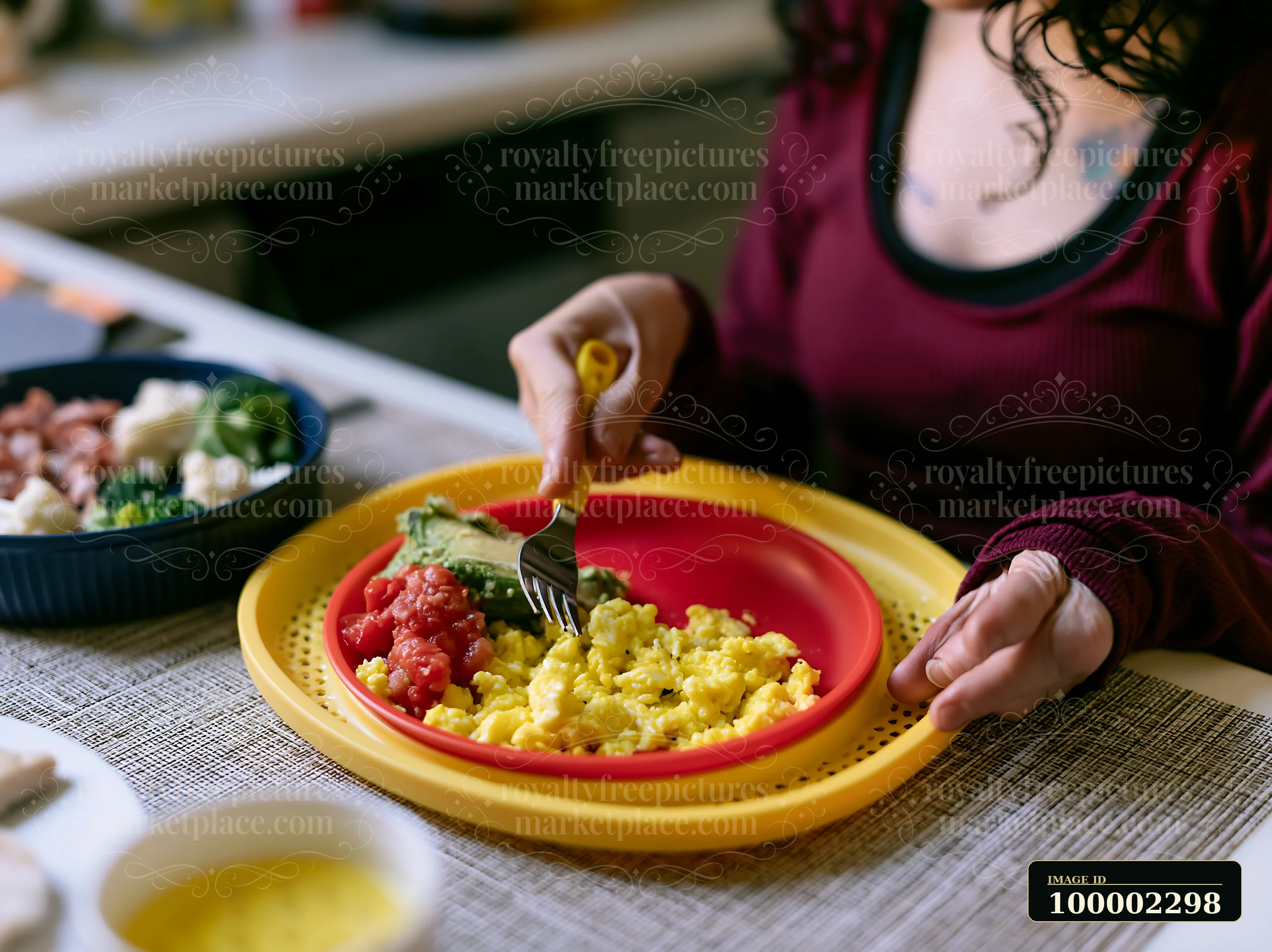 A person with limited hand mobility using utensils to eat scrambled eggs and vegetables from a red plate.