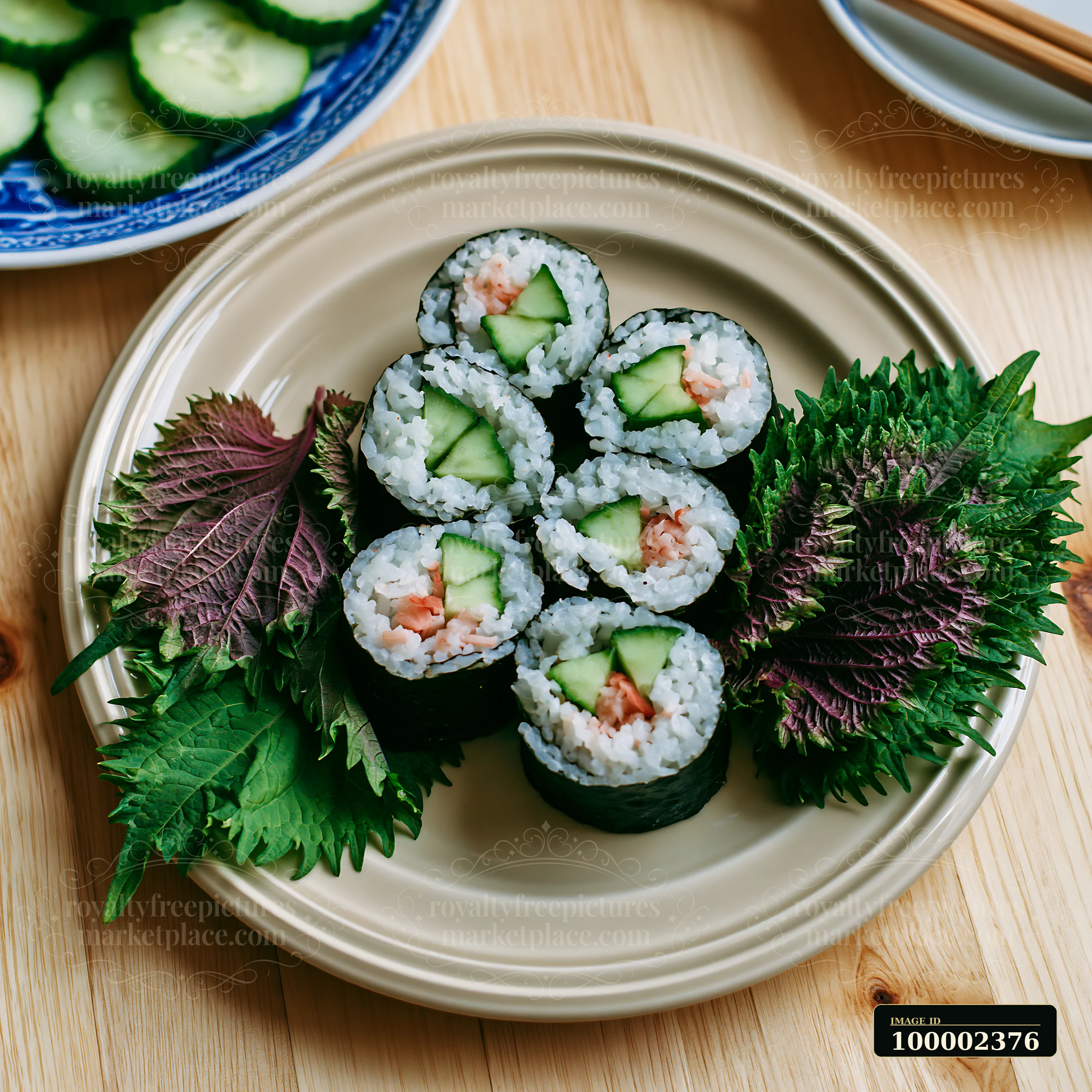 Six sushi rolls are arranged on a beige plate, surrounded by vibrant shiso leaves and cucumber slices on a wooden table.