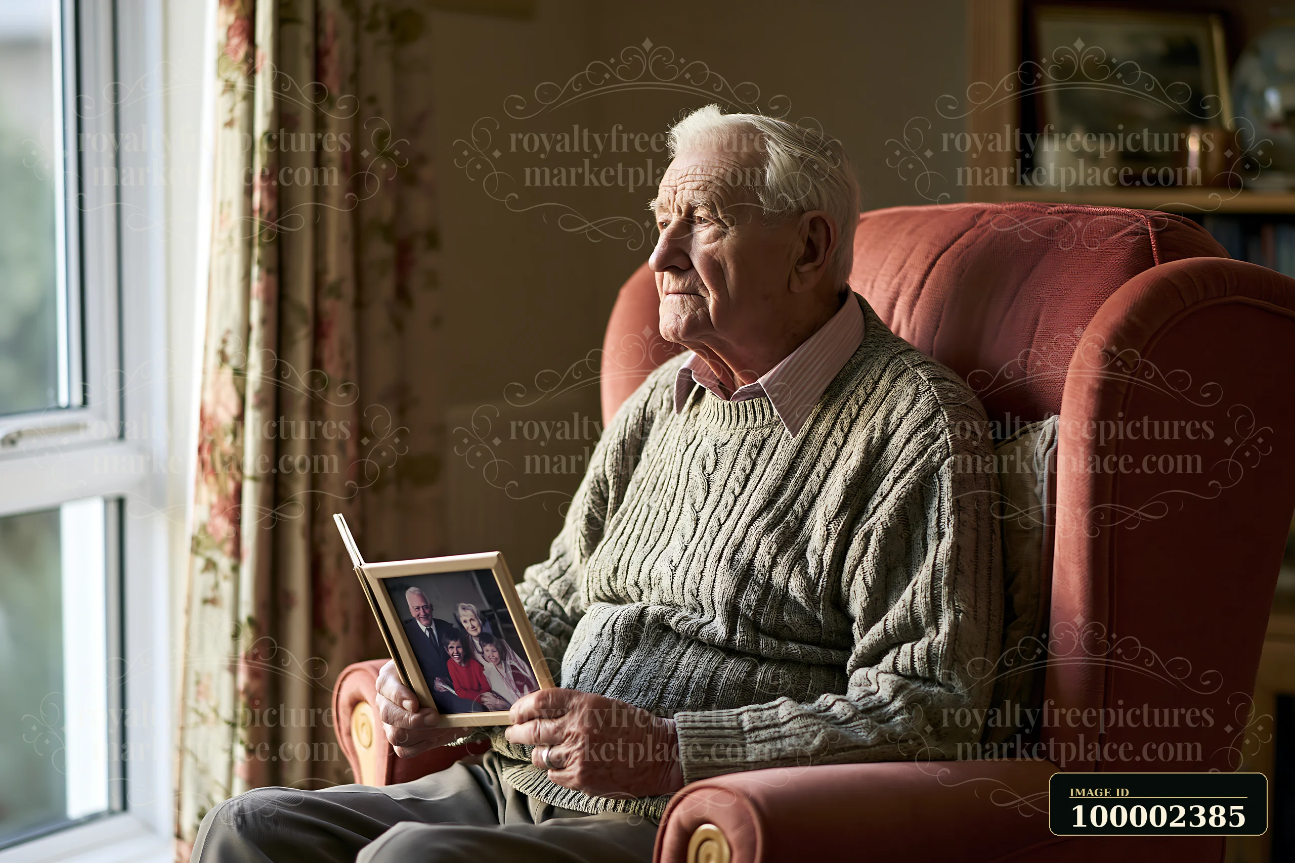 A senior man with Alzheimer's holding a photo of family sits in a cozy armchair by a window.