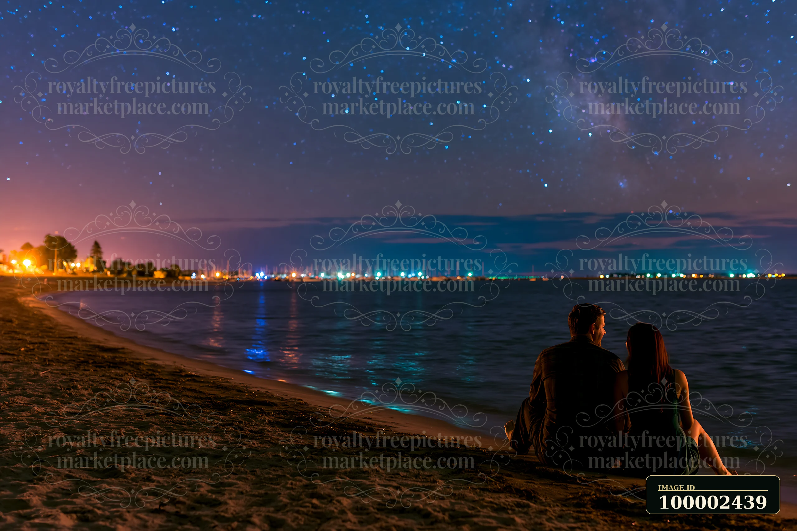 A couple on a beach under stars sits by the water, enjoying the view of the night sky filled with stars.