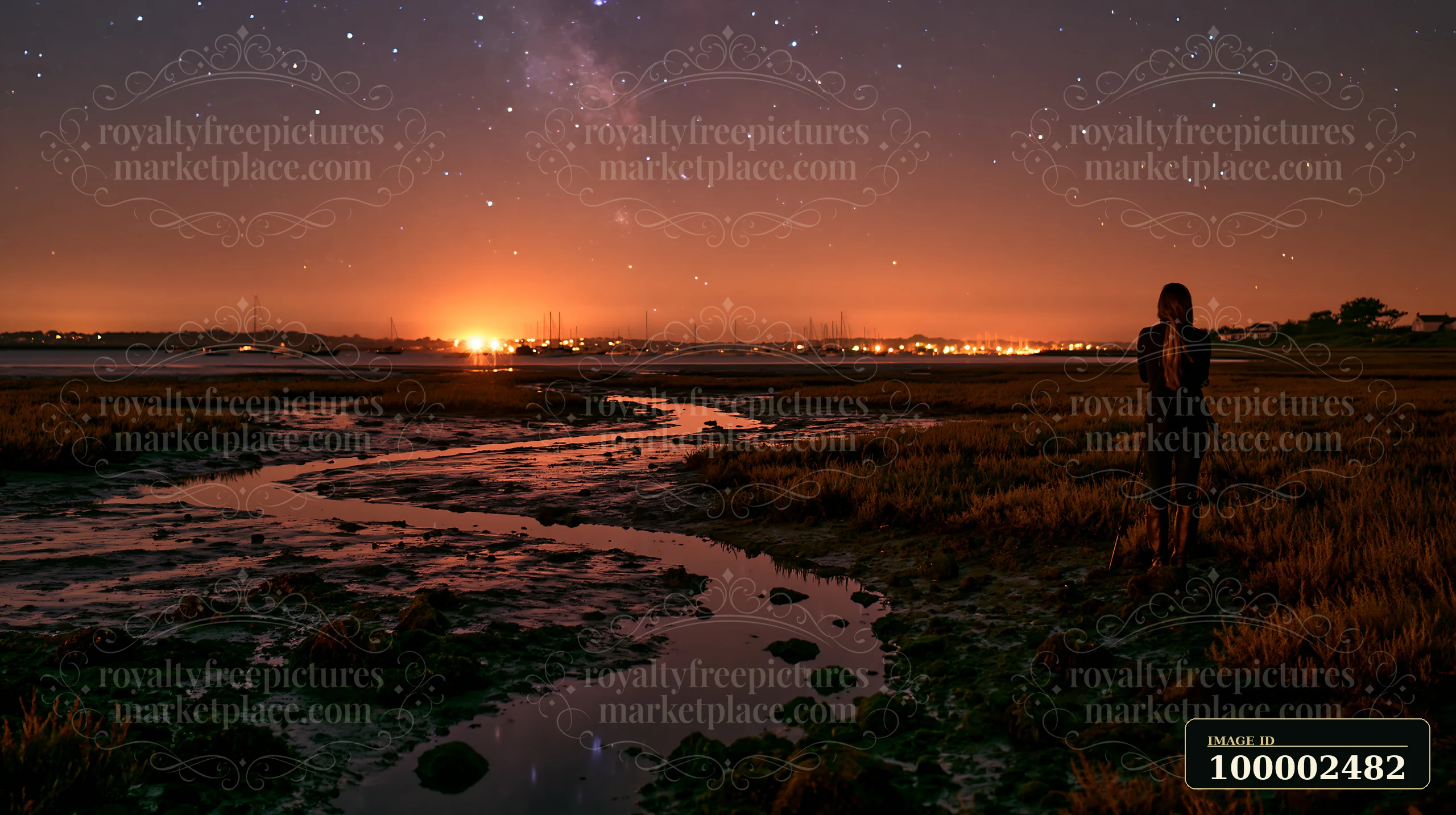 A person photographing stars at night stands near a river with low tide.