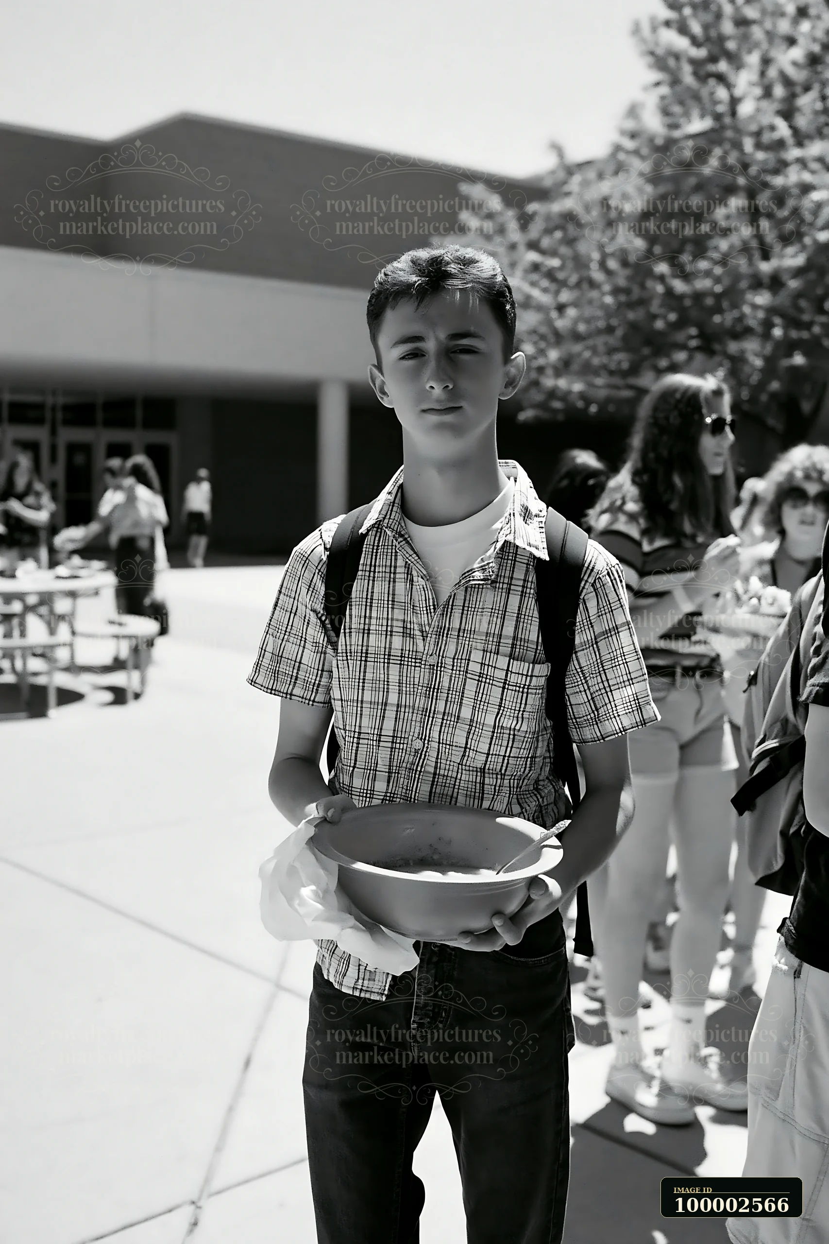 A young boy stands with a bowl in a line at a community potluck buffet during a sunny day outside a school.