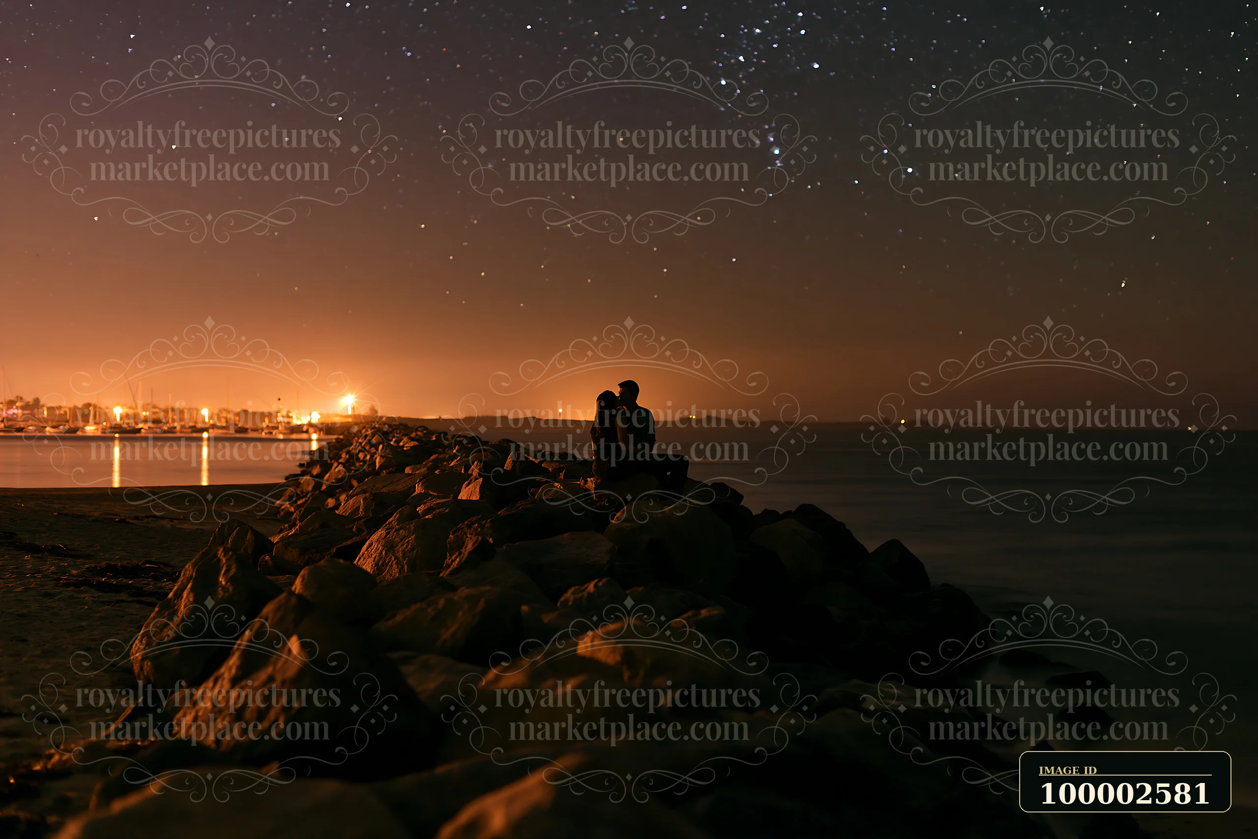 A couple shares a kiss silhouetted against a starry sky while seated on rocks at the beach.