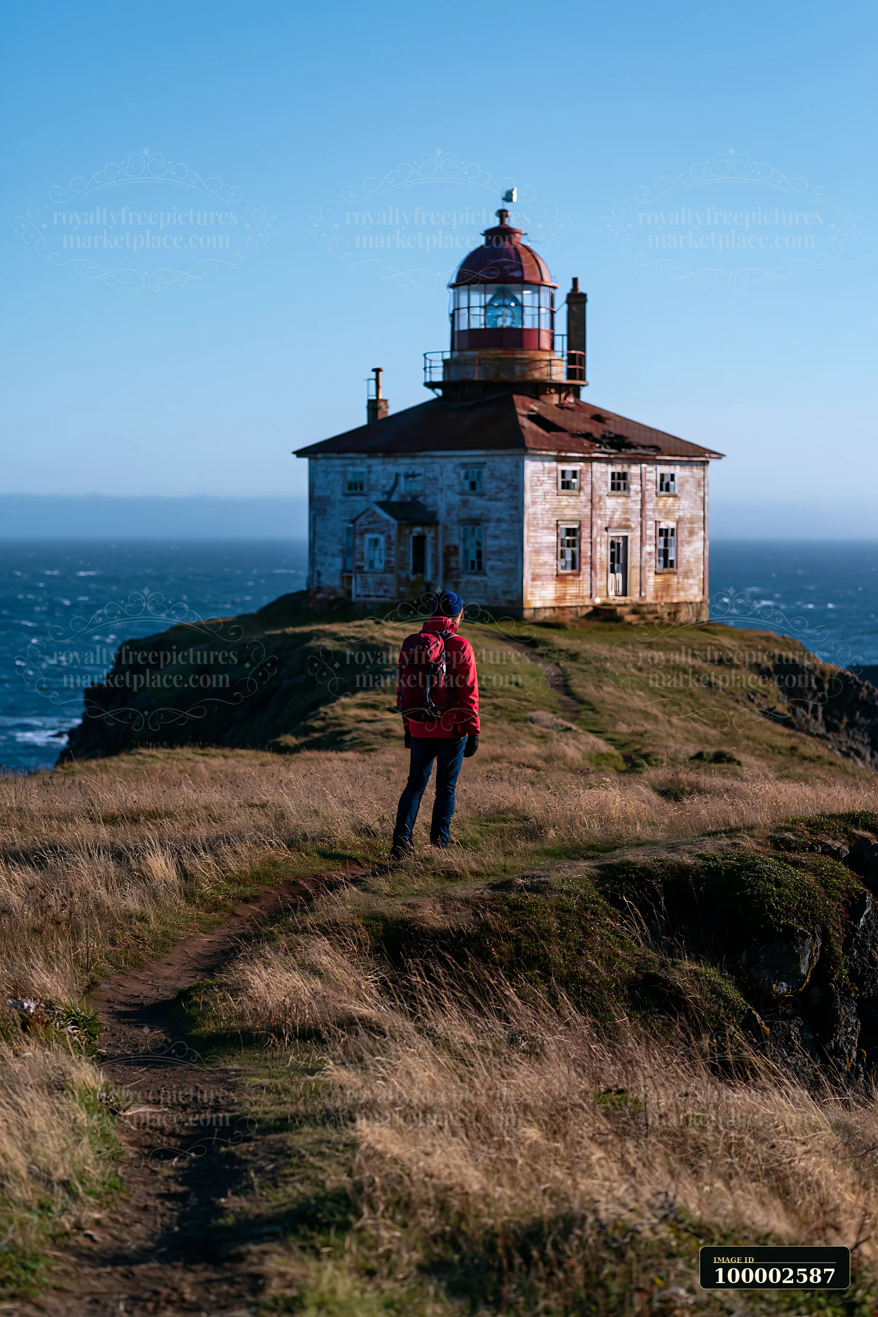 A person stands on a grassy path looking at an abandoned lighthouse on a remote coast.