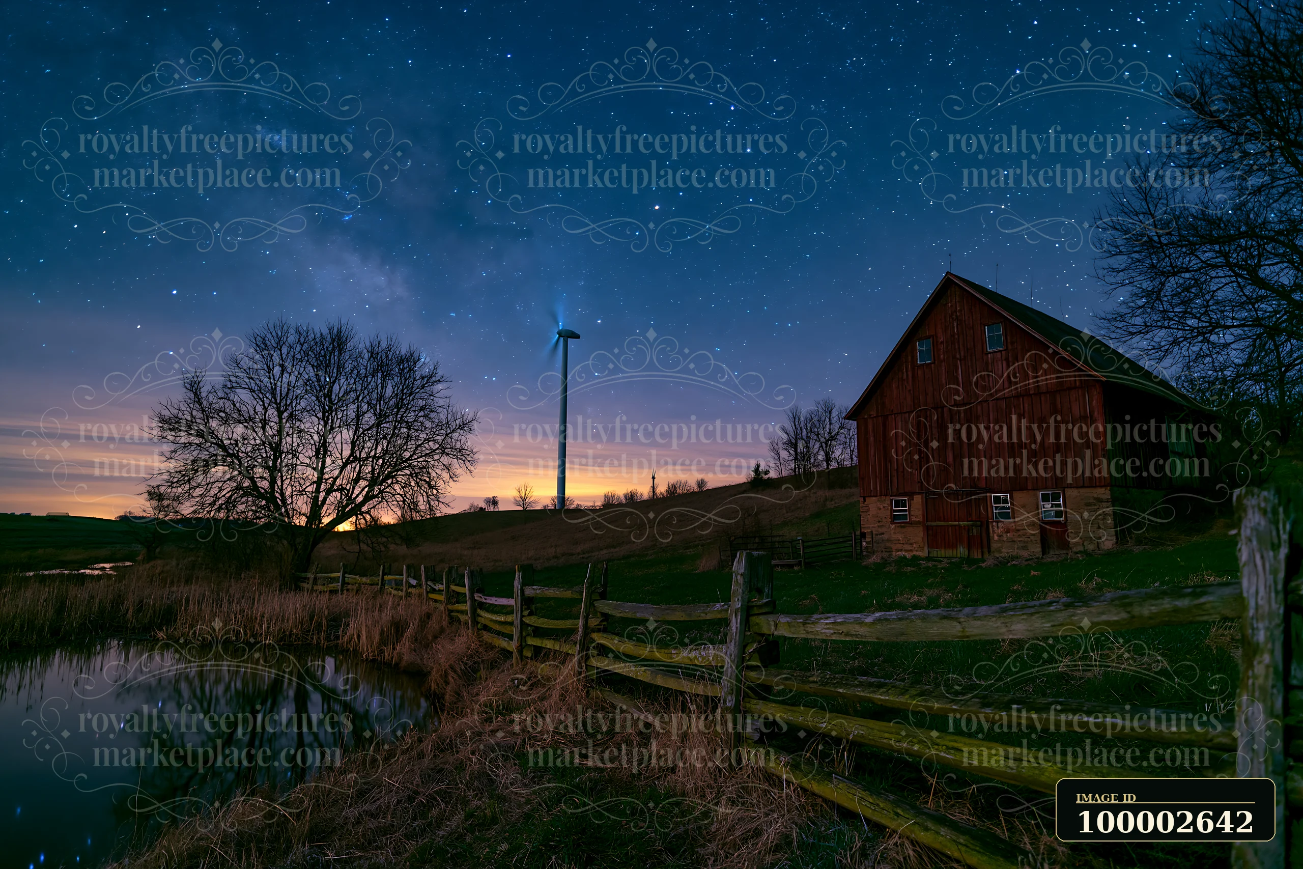 A wind turbine stands near a barn by a river under a starry sky.