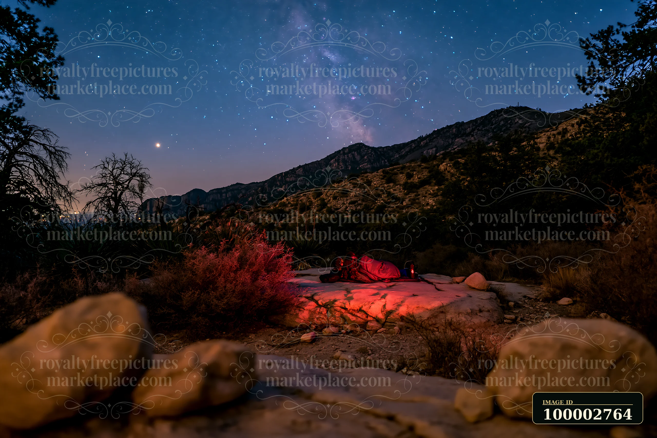 A couple stargazing in a national park sits on rocky terrain under a star-filled sky.