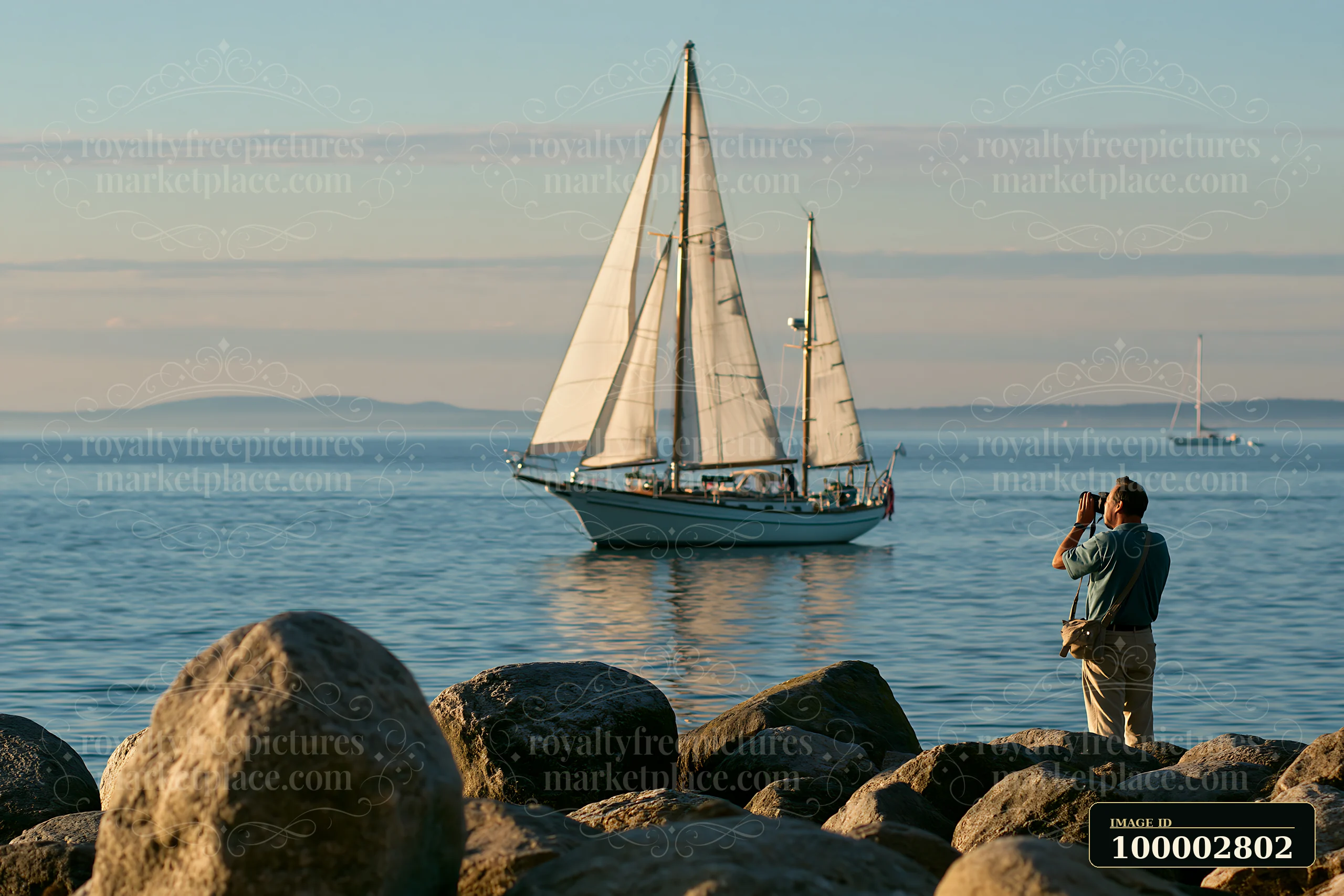 A photographer photographing a sailboat at sea stands on the shore among rocky formations.