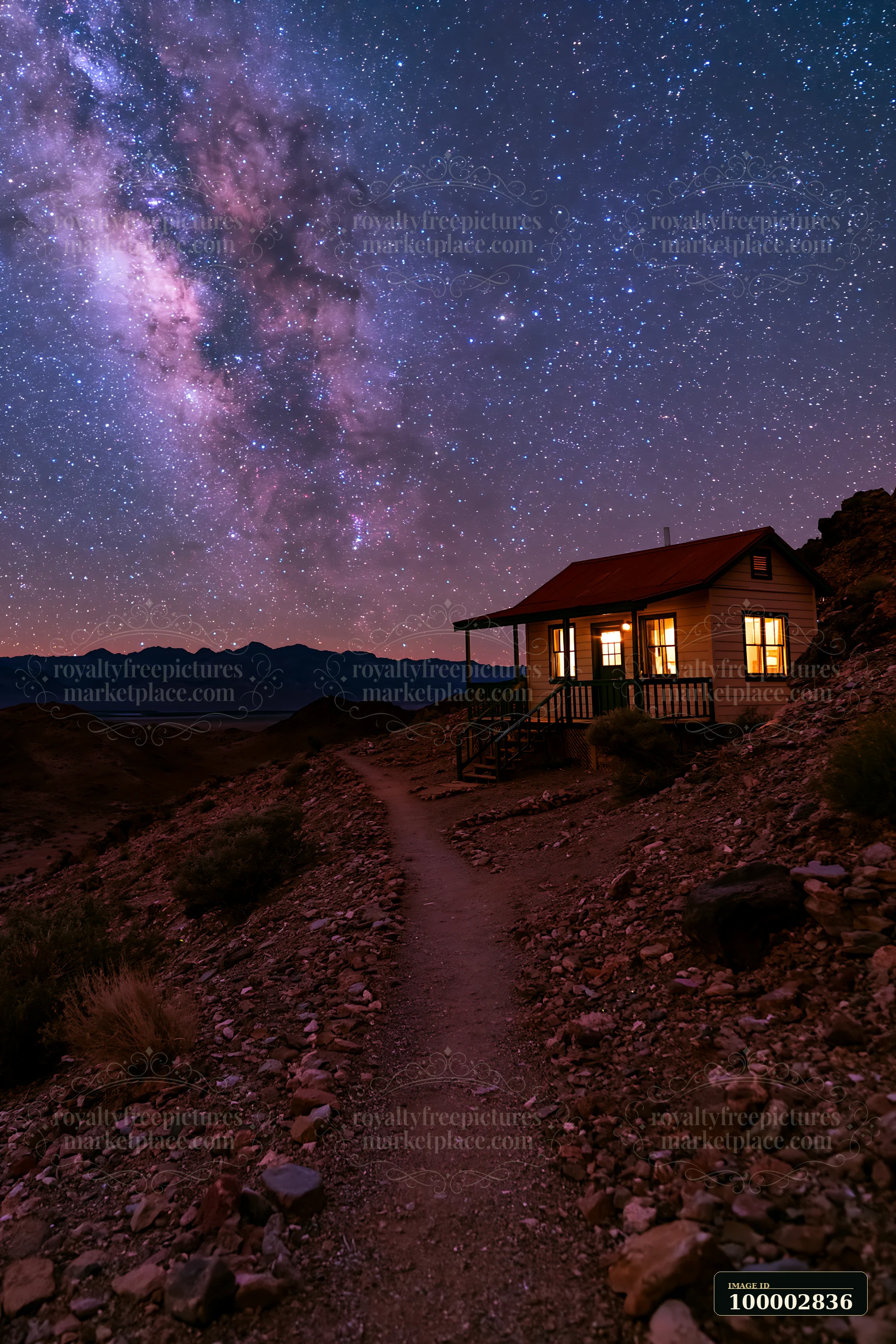 A remote house sits on a rocky path surrounded by mountains, illuminated by the warm glow of its windows.