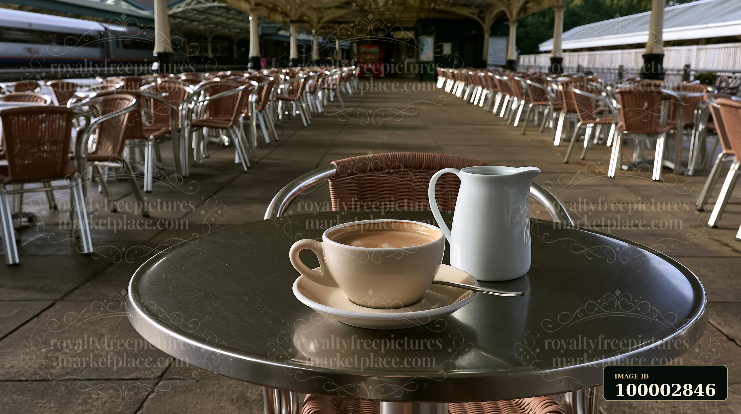 An outdoor cafe table features a latte and a milk jug, set in a spacious area with empty chairs and a roofed seating arrangement.