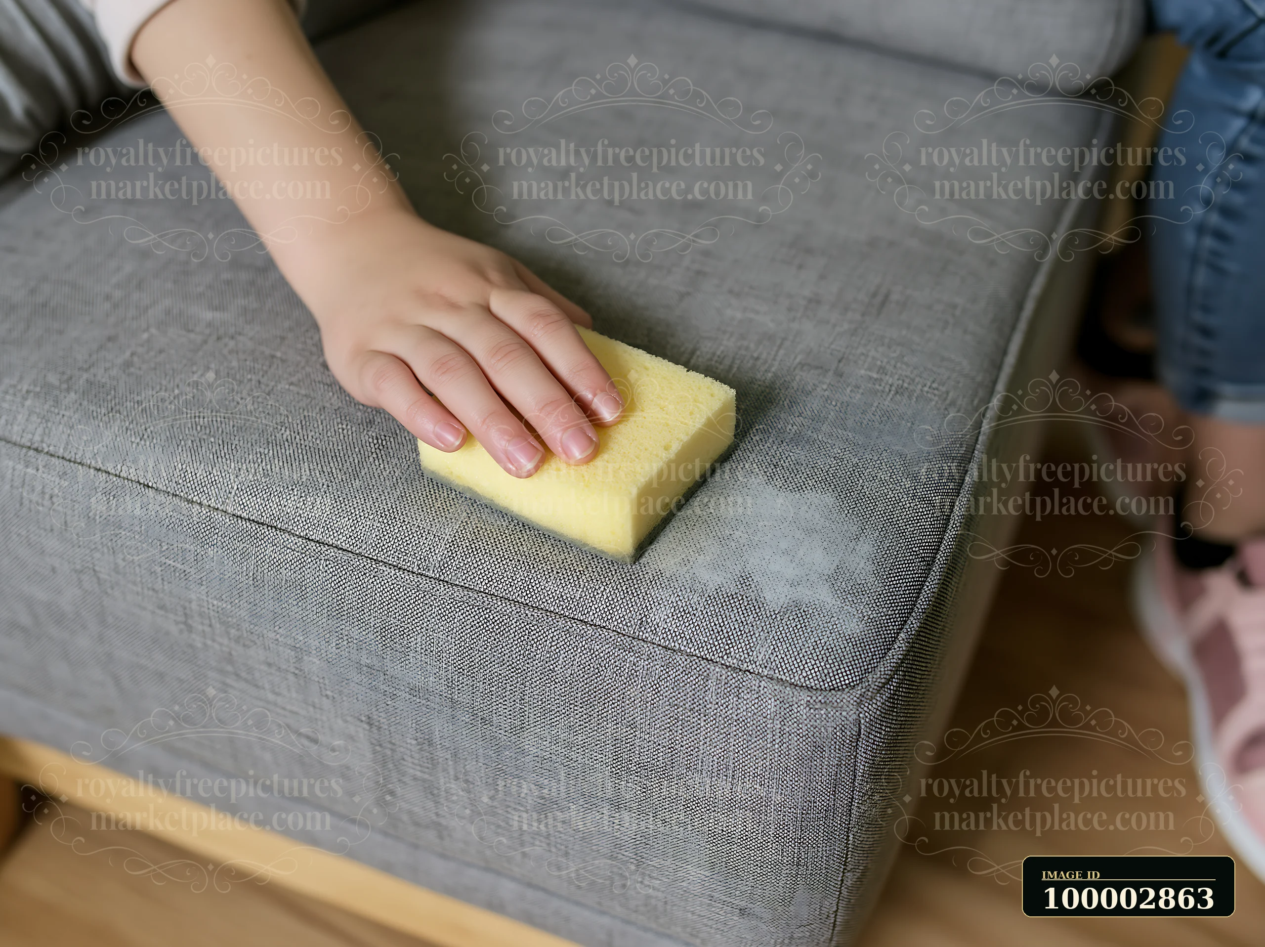 A person uses a yellow sponge to remove a stain from a gray upholstered couch in a living room setting.