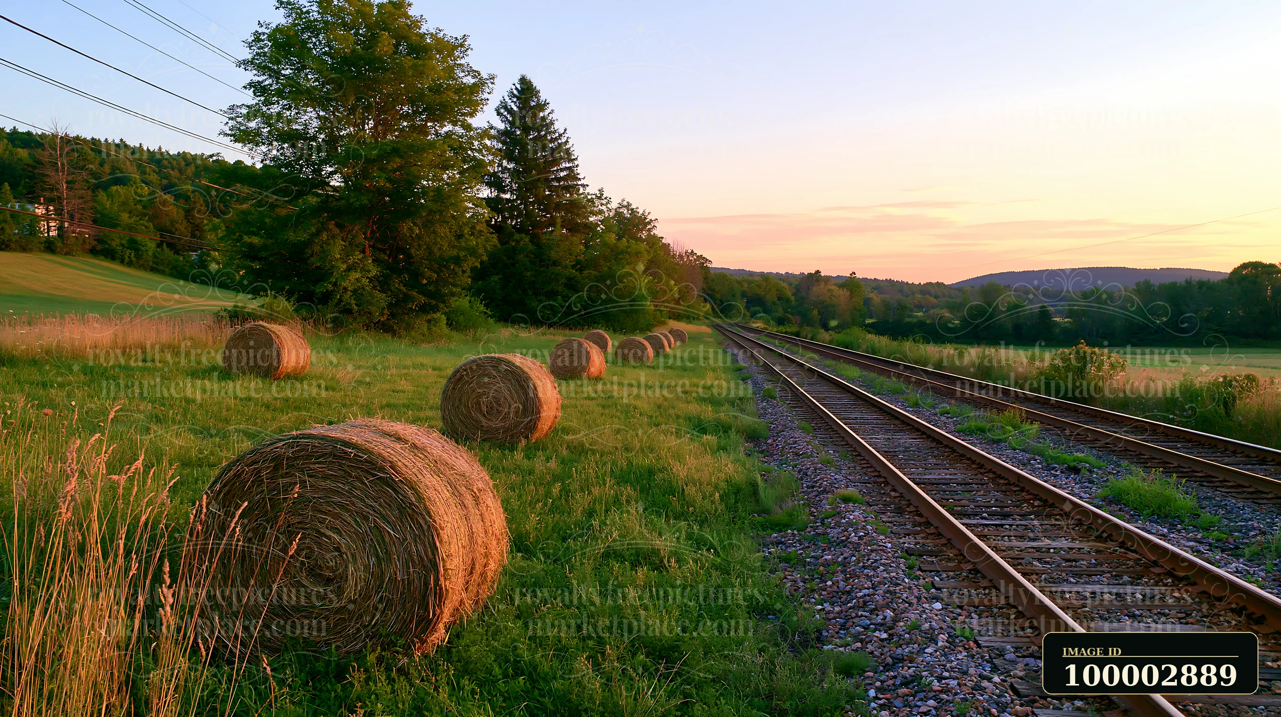 Bales of hay line a meadow alongside railway tracks near the sunset.