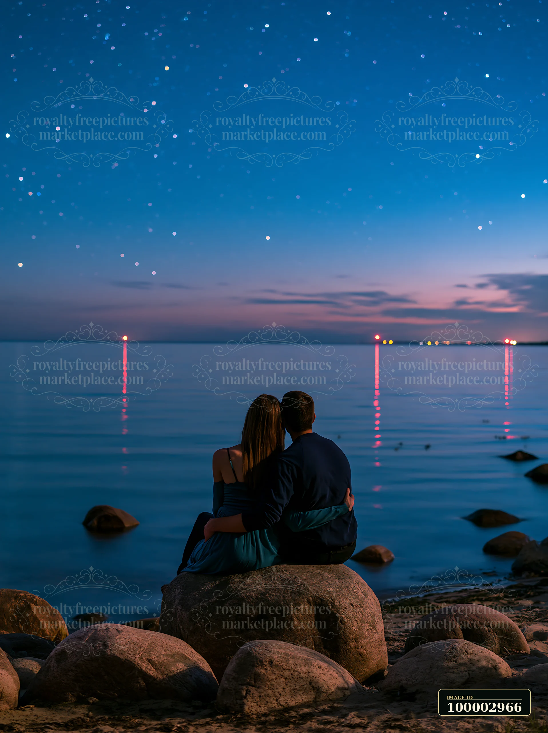 A couple sits on a rock by the water, looking at the stars above.