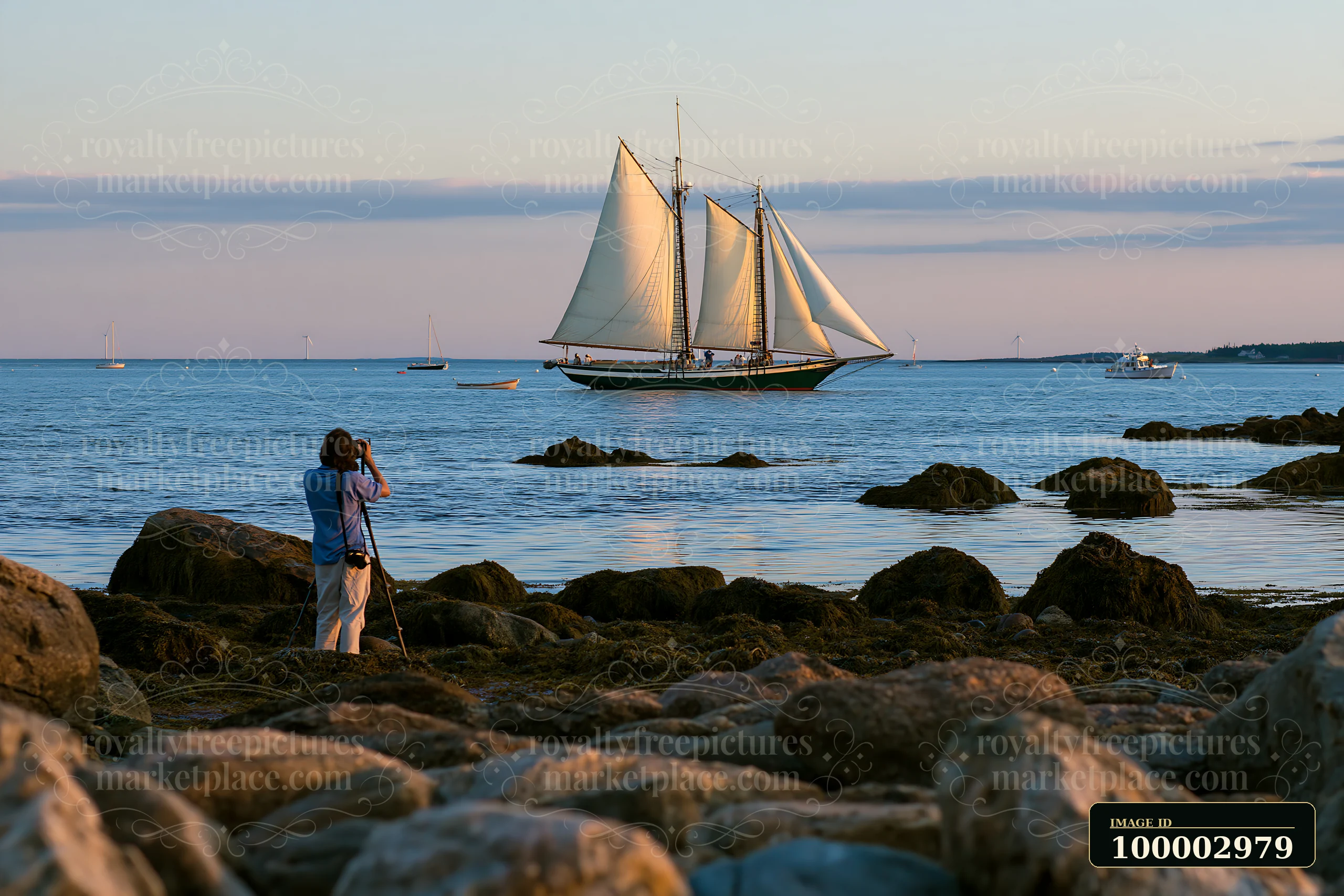 A photographer photographing a sailboat at sea stands on rocky shore.