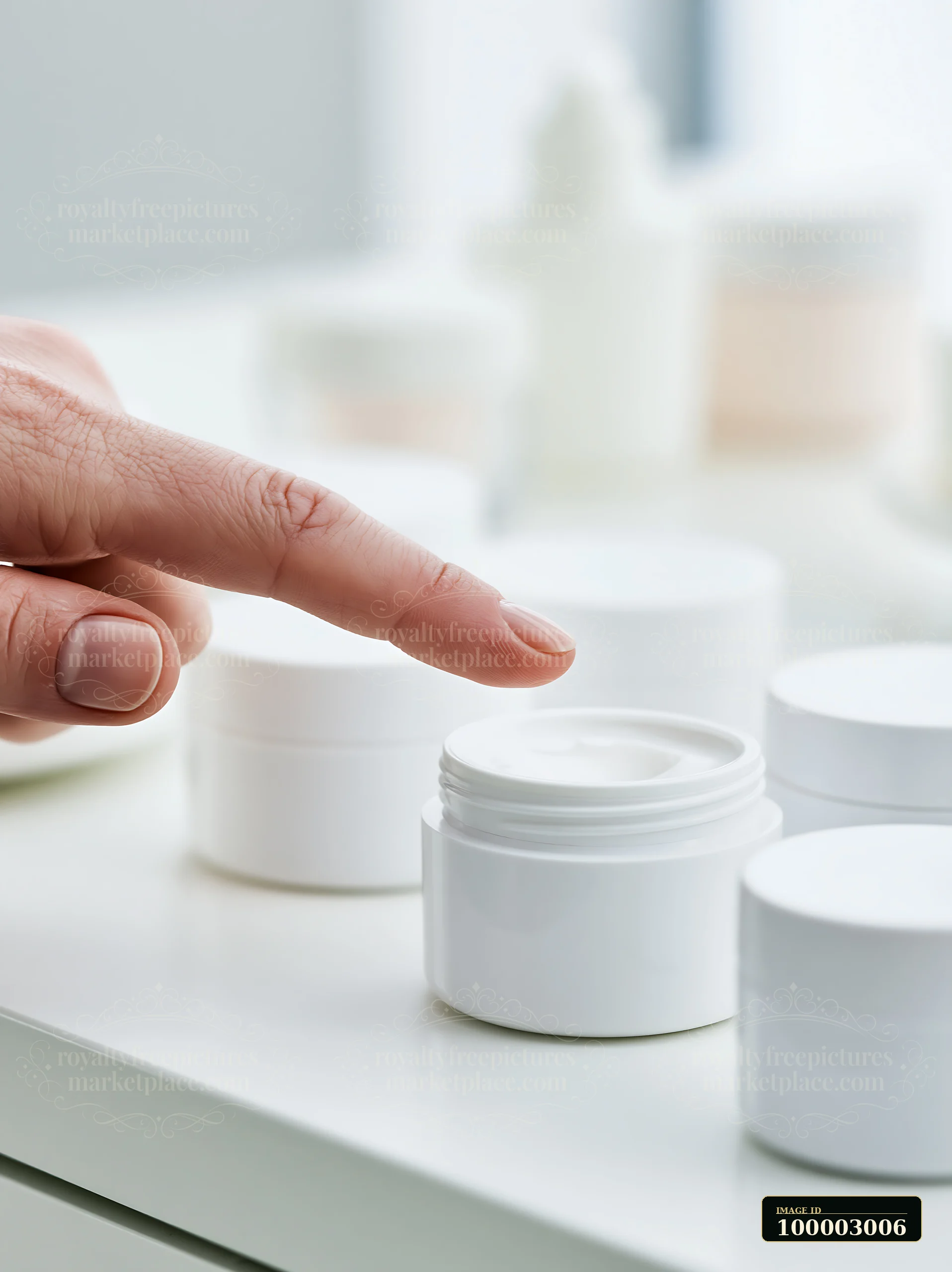 A hand reaches towards a small container of cream on a white tabletop surrounded by unbranded cosmetic containers.