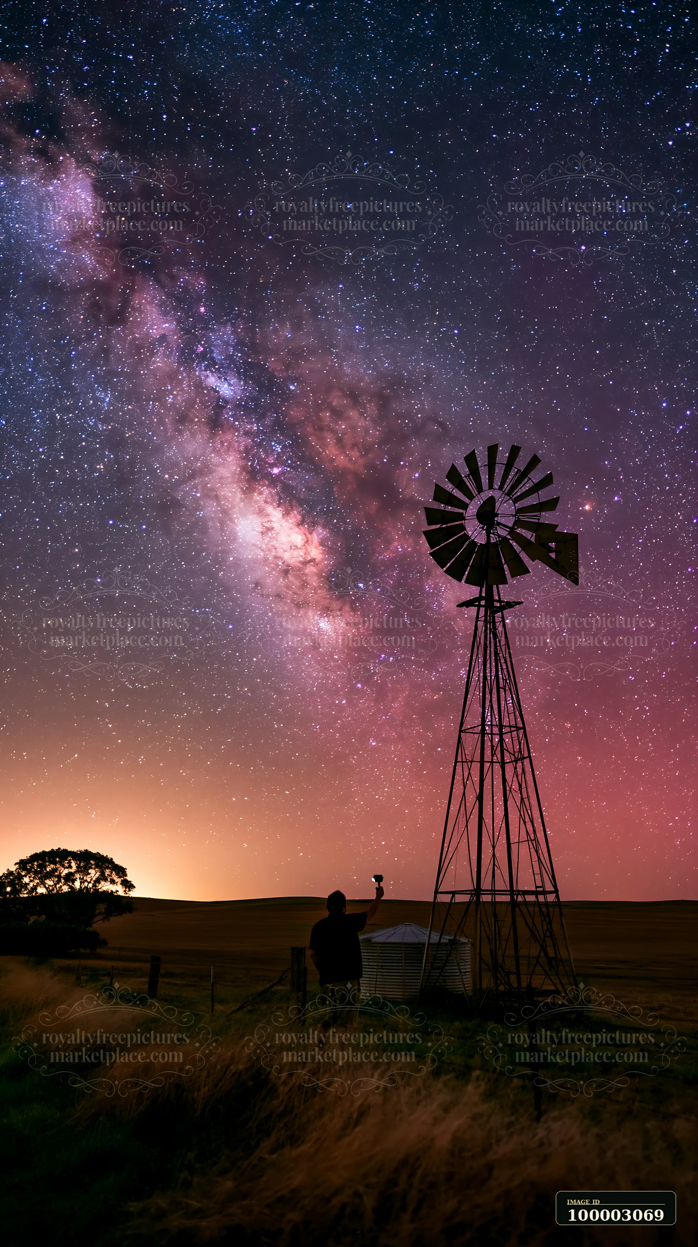 A person using camera under Milky Way stands near a windmill in a field.